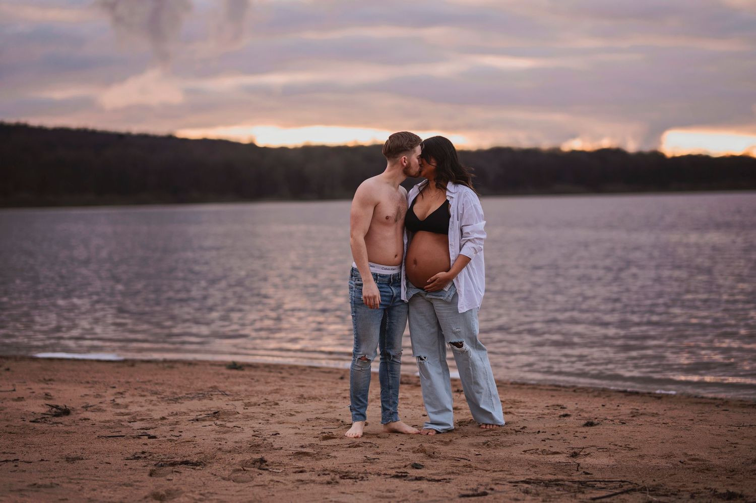 A romantic couple embraces on a beach shore at sunset with a lake backdrop.