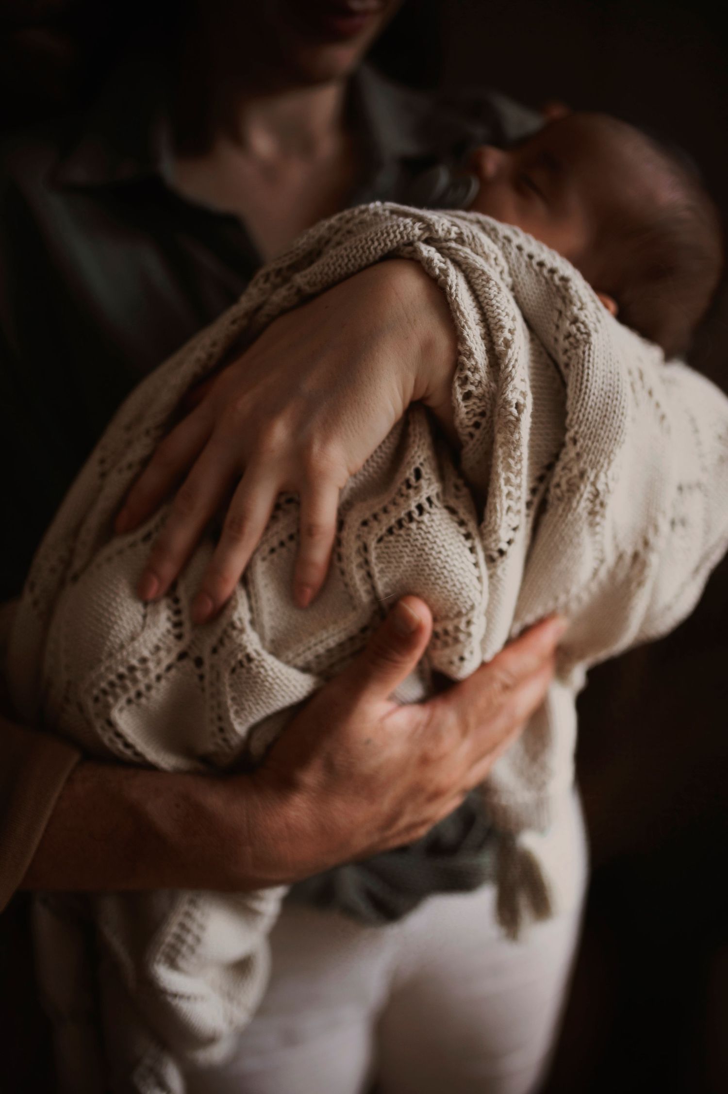 Infant wrapped in cozy knit blanket being tenderly held against caregiver in moody lighting.