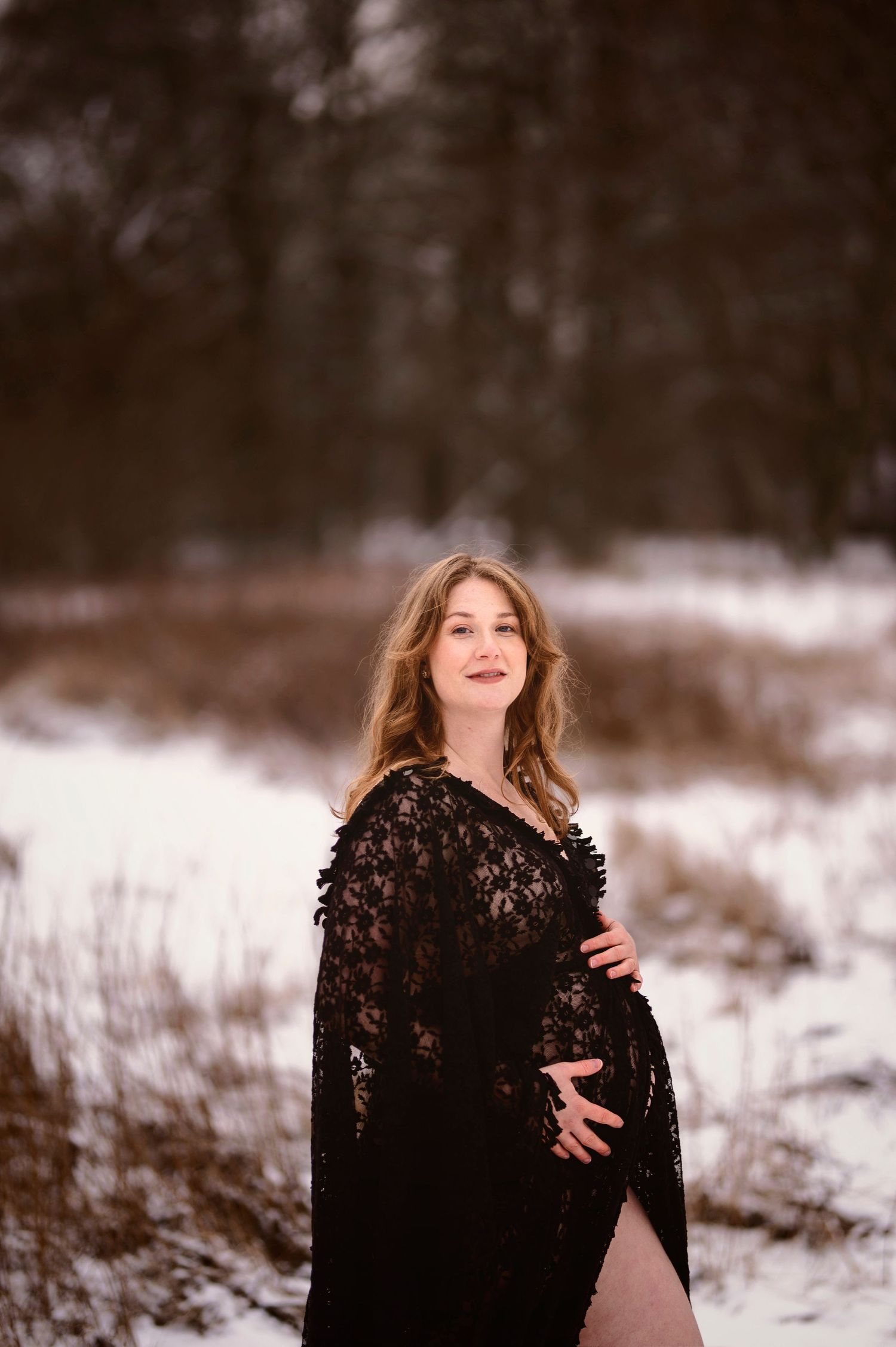 Gravidfotografering i Skåne med gravid kvinna i svart spetsklänning i snöig vinterskog.