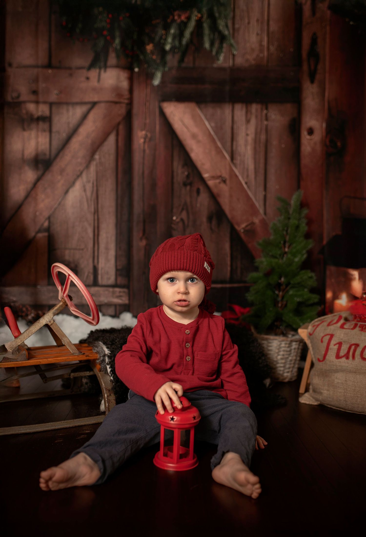 A toddler in red outfit and knit hat sits barefoot holding a red lantern in a festive Christmas barn setting.