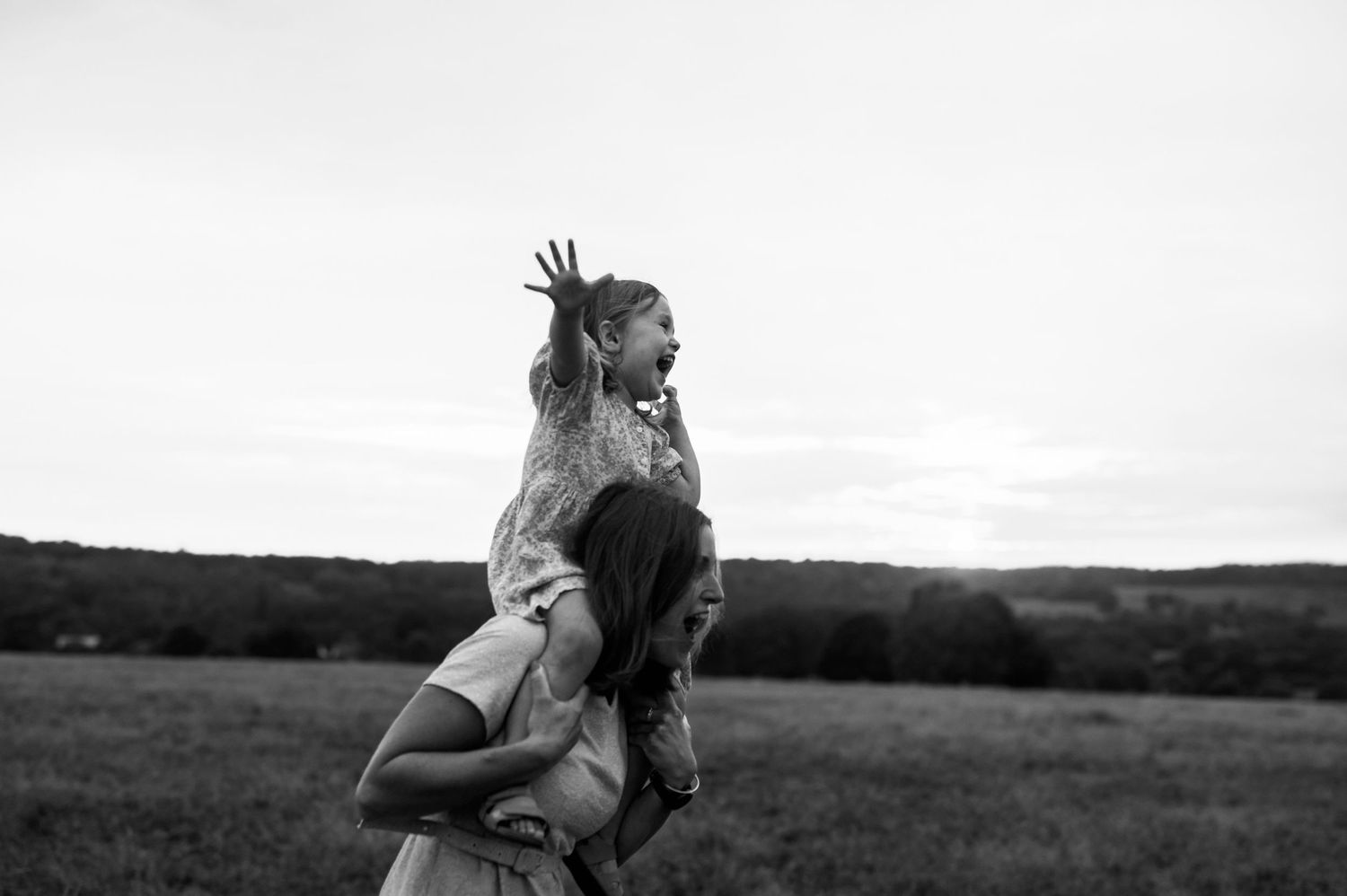 Familjefotografering i Skåne med barn som sitter på mammas axlar i svartvitt ljus i öppet fält.