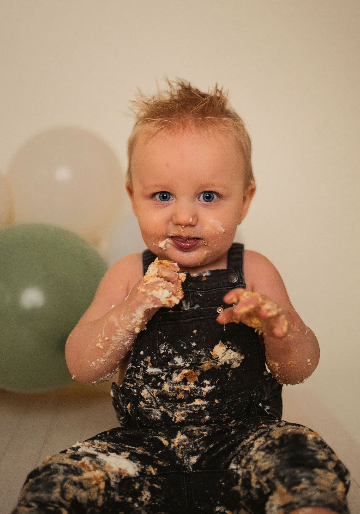A joyful cake smash portrait with frosting-covered overalls and balloon decorations.