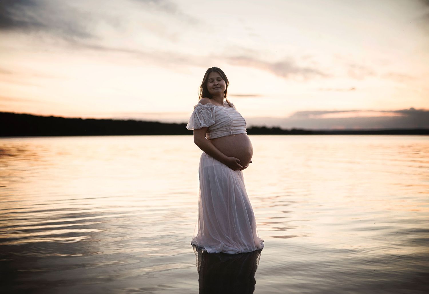 Maternity photo showing woman in flowing dress standing in water at dusk.