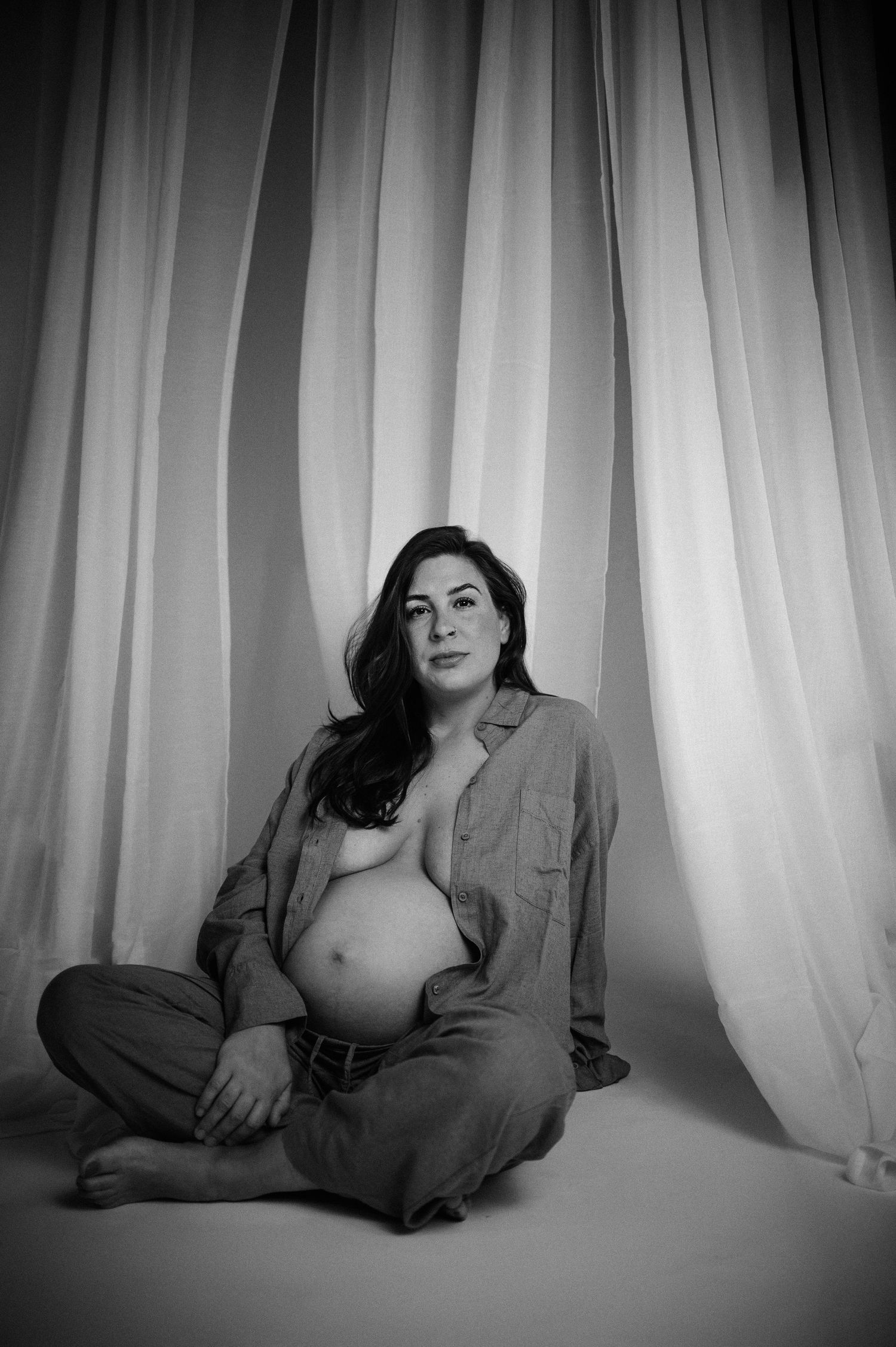 Black and white maternity photo of pregnant woman sitting cross-legged before flowing white curtains.