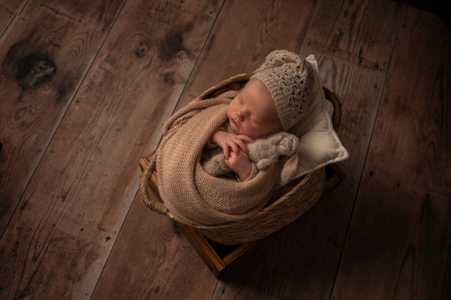 Newborn baby sleeping peacefully in a wooden basket, wrapped in a beige knit blanket and wearing a matching bear ear hat.