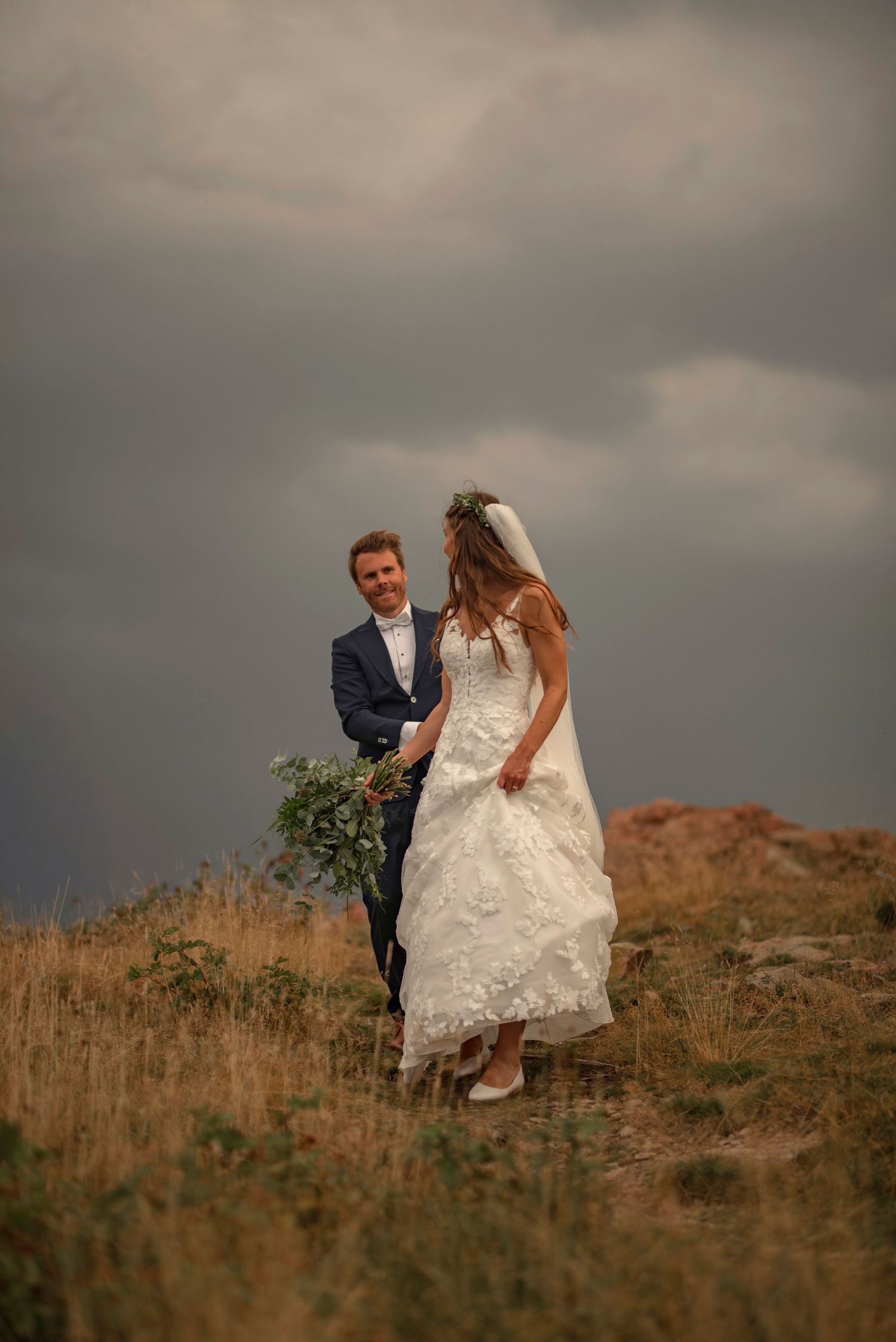 Elopement at Hovs Hallar in Skåne, Sweden with bride and groom walking on hilltop under dramatic stormy sky, bride in lace dress and veil.