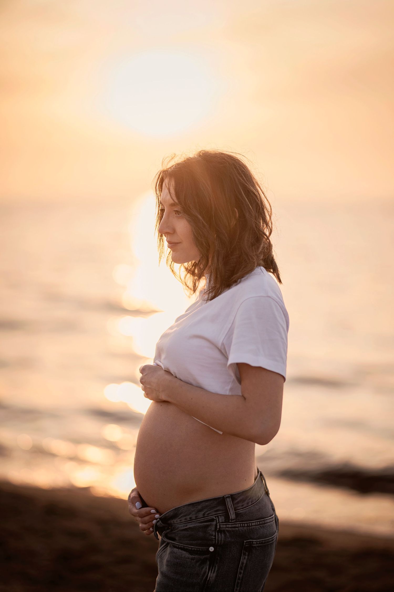Gravidfotografering i Skåne med gravid kvinna vid havet i solnedgång i mjukt kvällsljus.