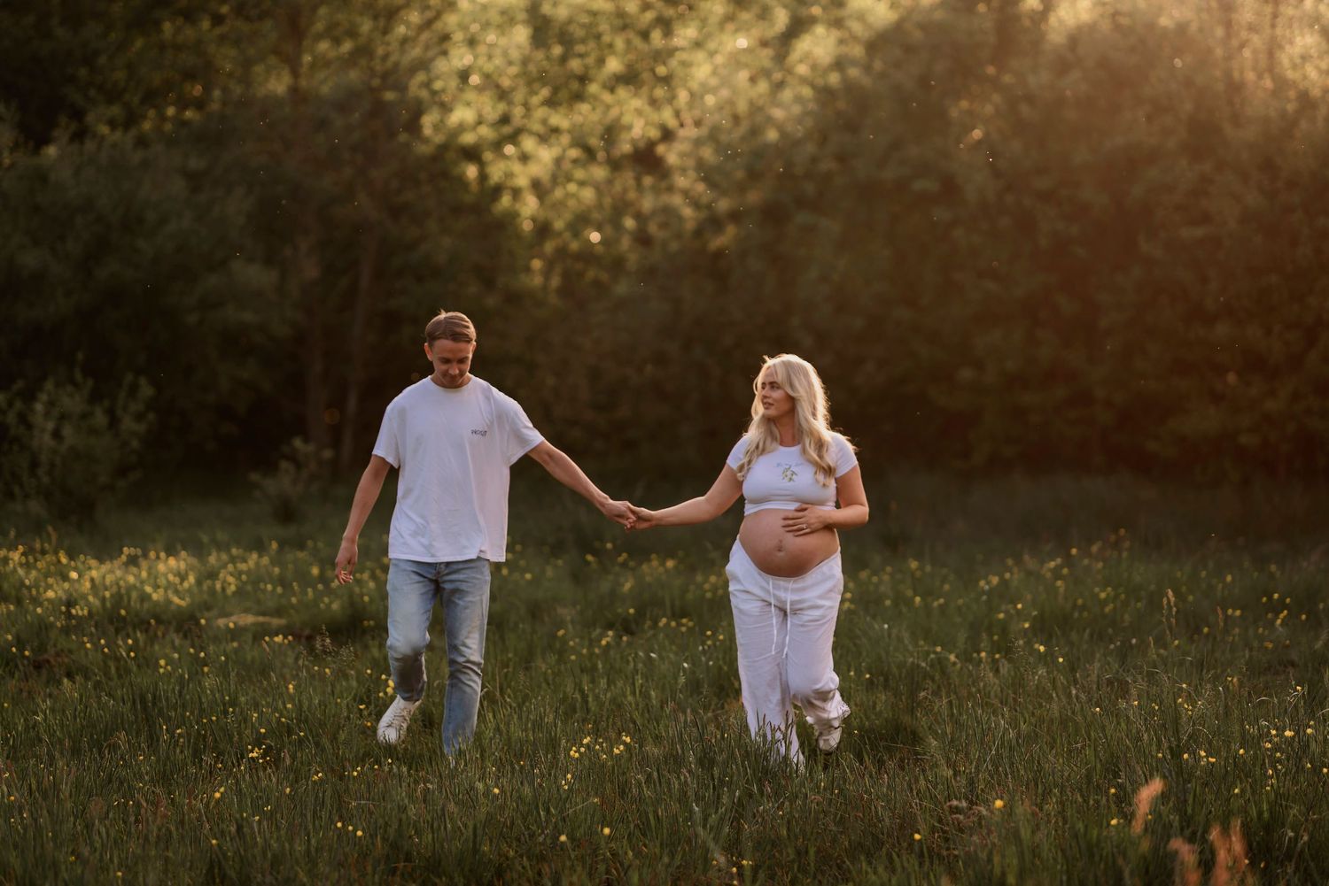 Gravidfotografering i Skåne med blivande föräldrar som går hand i hand i en solbelyst äng.