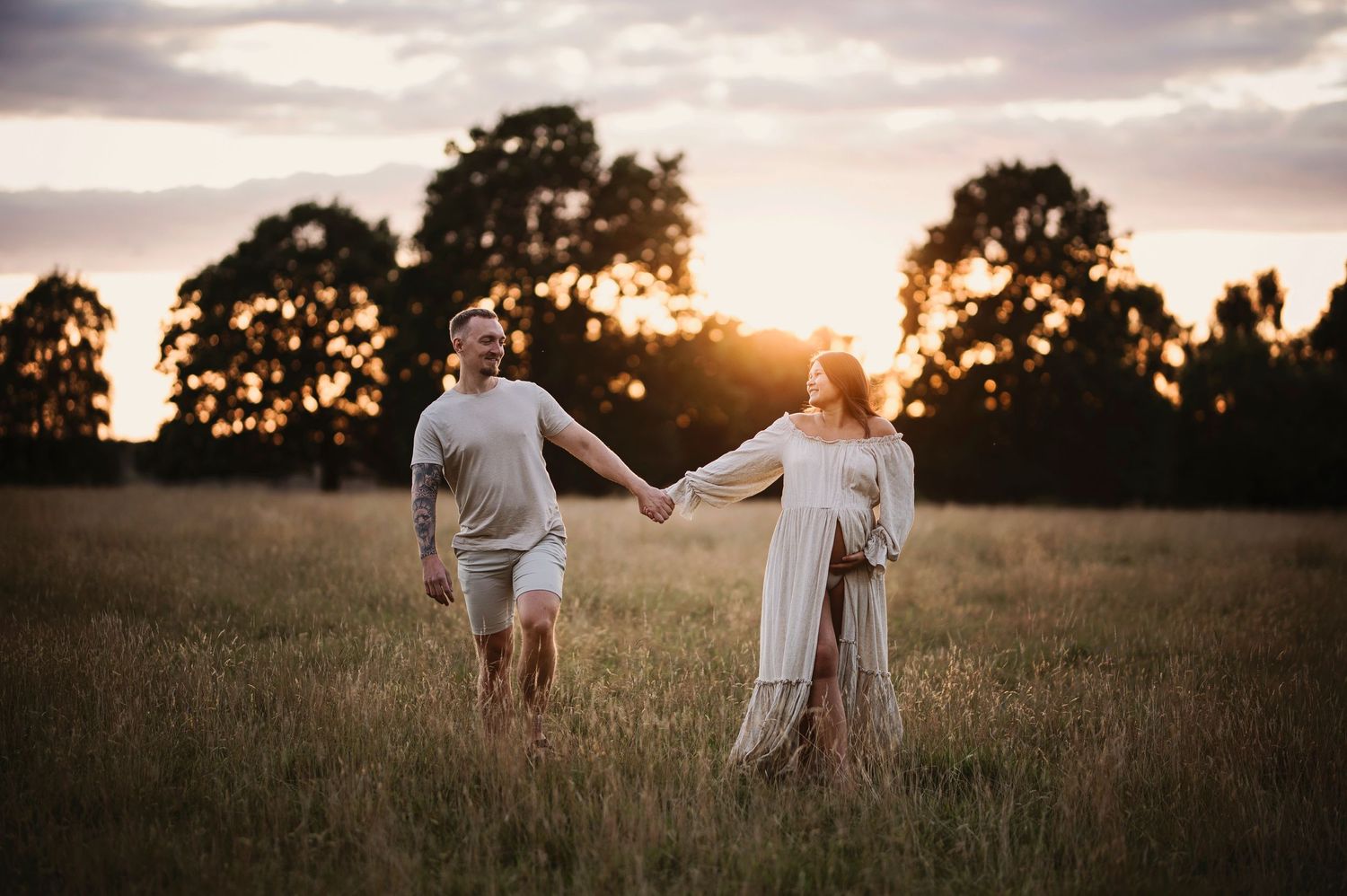 A couple holding hands while walking through a sunlit field at dusk.