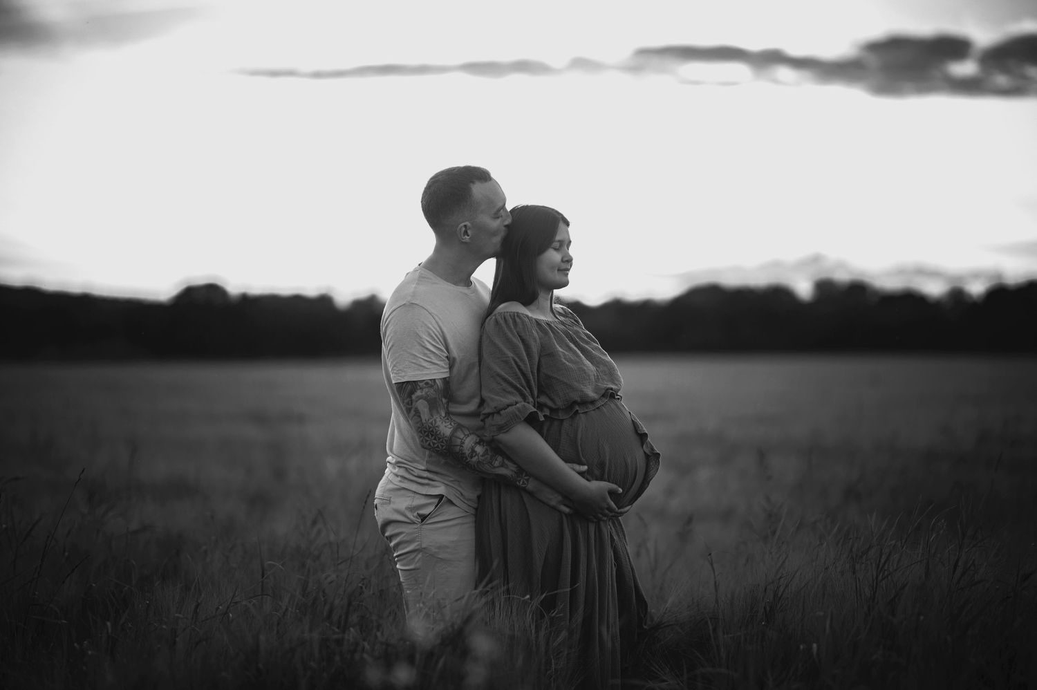 A black and white maternity photo of a couple embracing in a peaceful meadow at dusk.