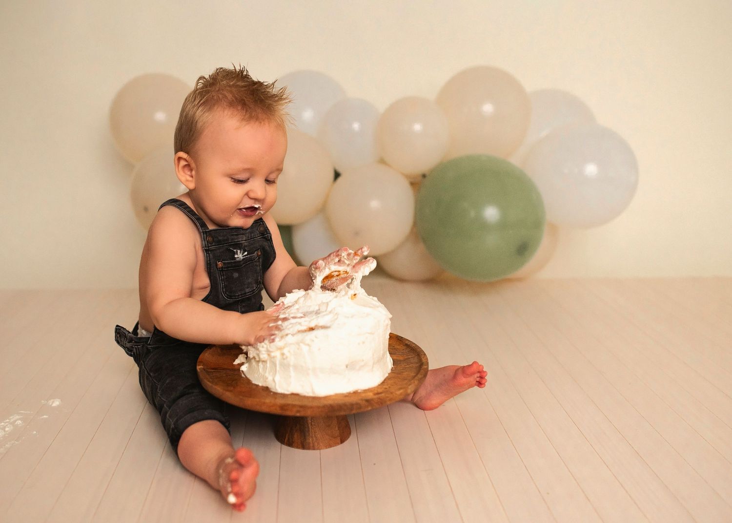 A cake smash session showing small hands reaching for white frosting on a wooden cake stand.