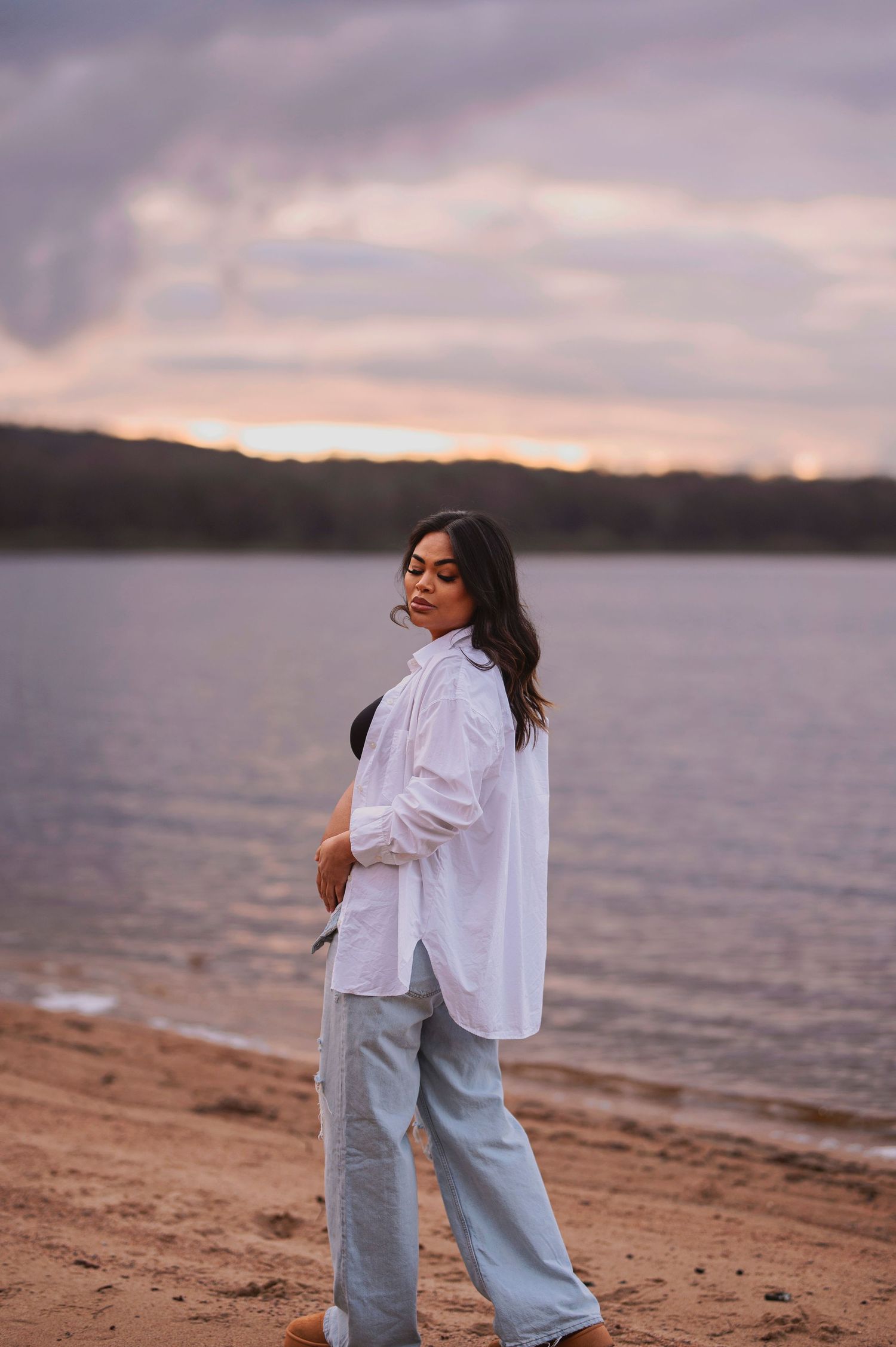 Maternity photo featuring woman in white shirt and jeans on lakeside during sunset.