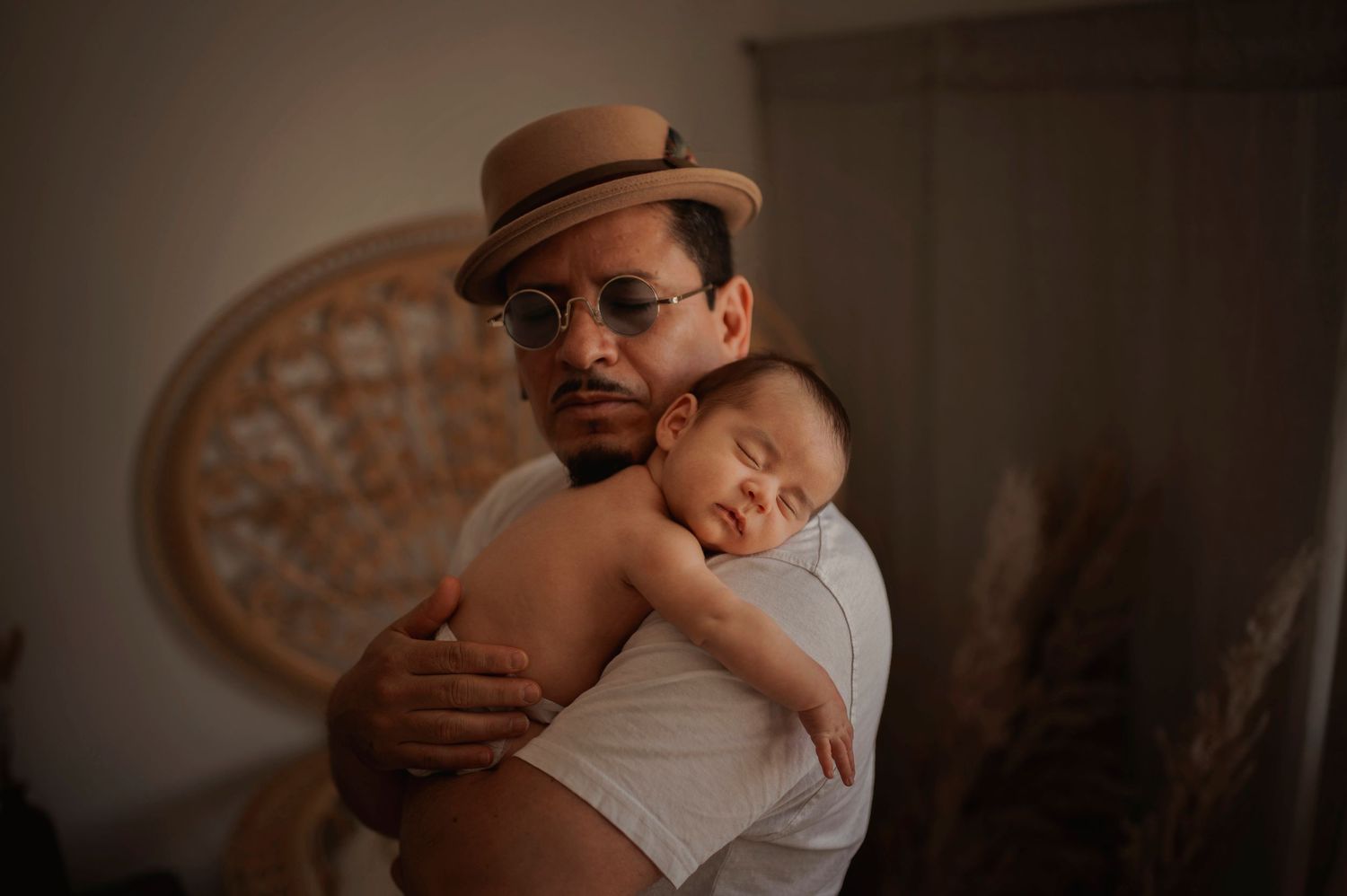 Tender moment of sleeping baby resting on caregiver's shoulder wearing brown fedora hat.