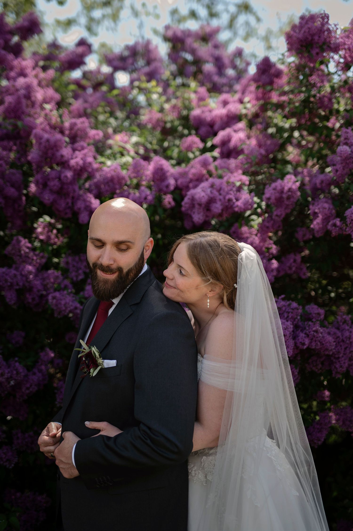 Bride leaning on groom's back in front of blooming purple lilac bushes in romantic wedding portrait.