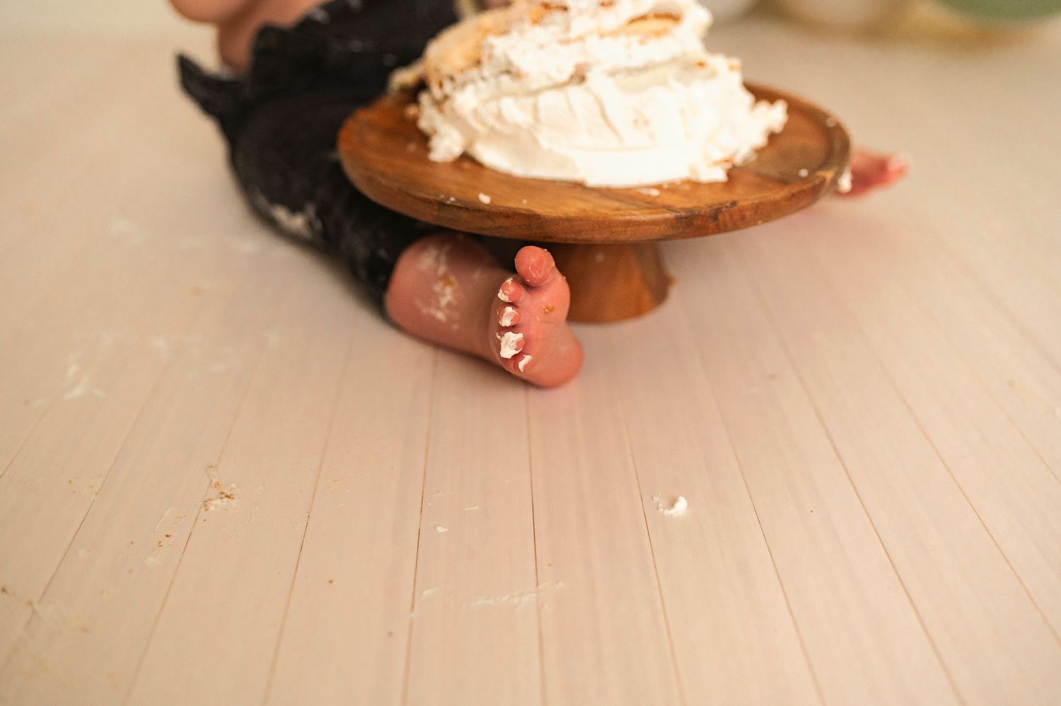 Close-up of tiny feet next to a wooden cake stand with white frosting.