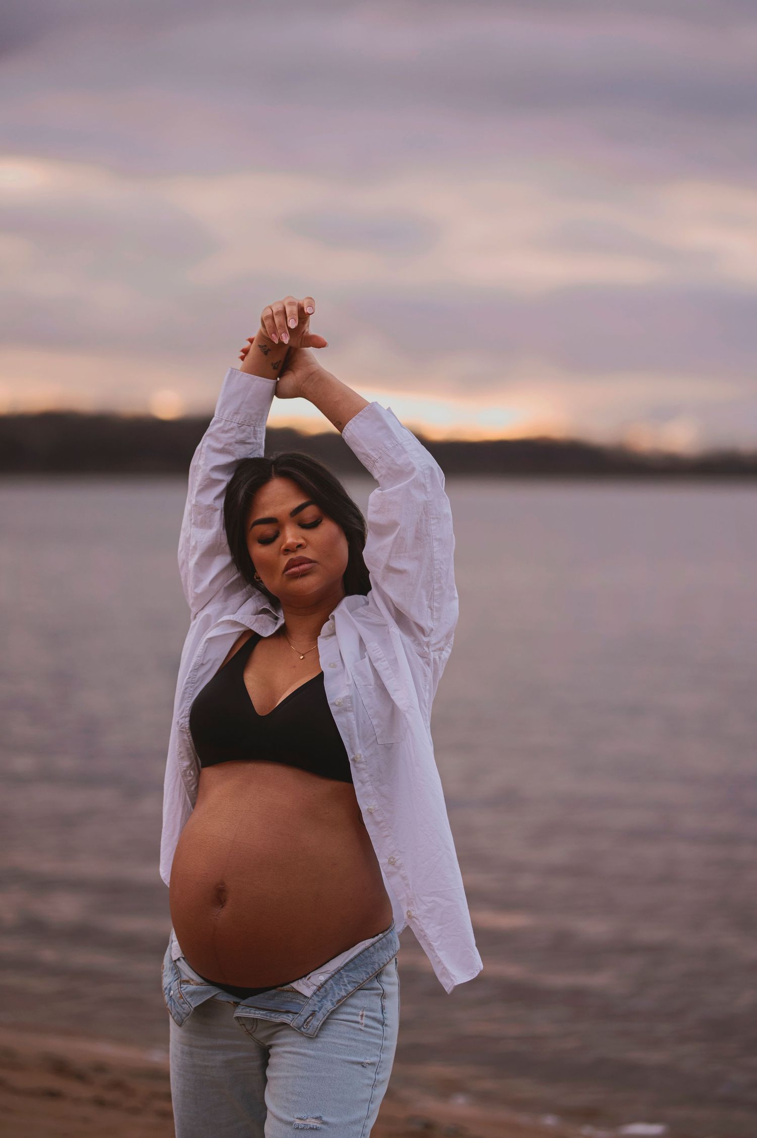 Pregnant woman in white shirt strikes artistic pose on beach at sunset.