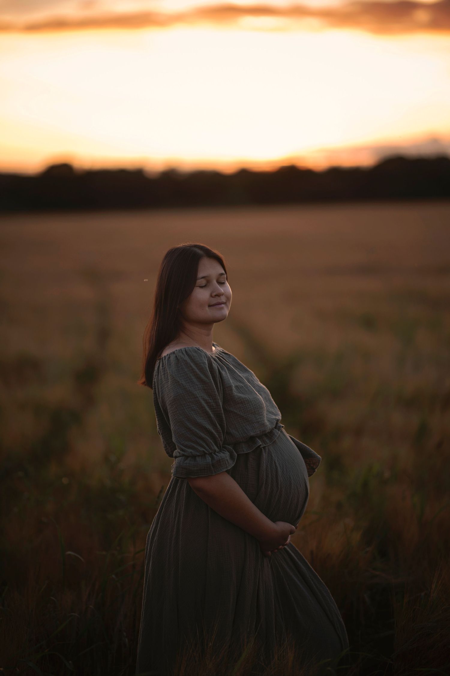 A maternity portrait of an expectant mother in a grey dress standing in a golden field at sunset.