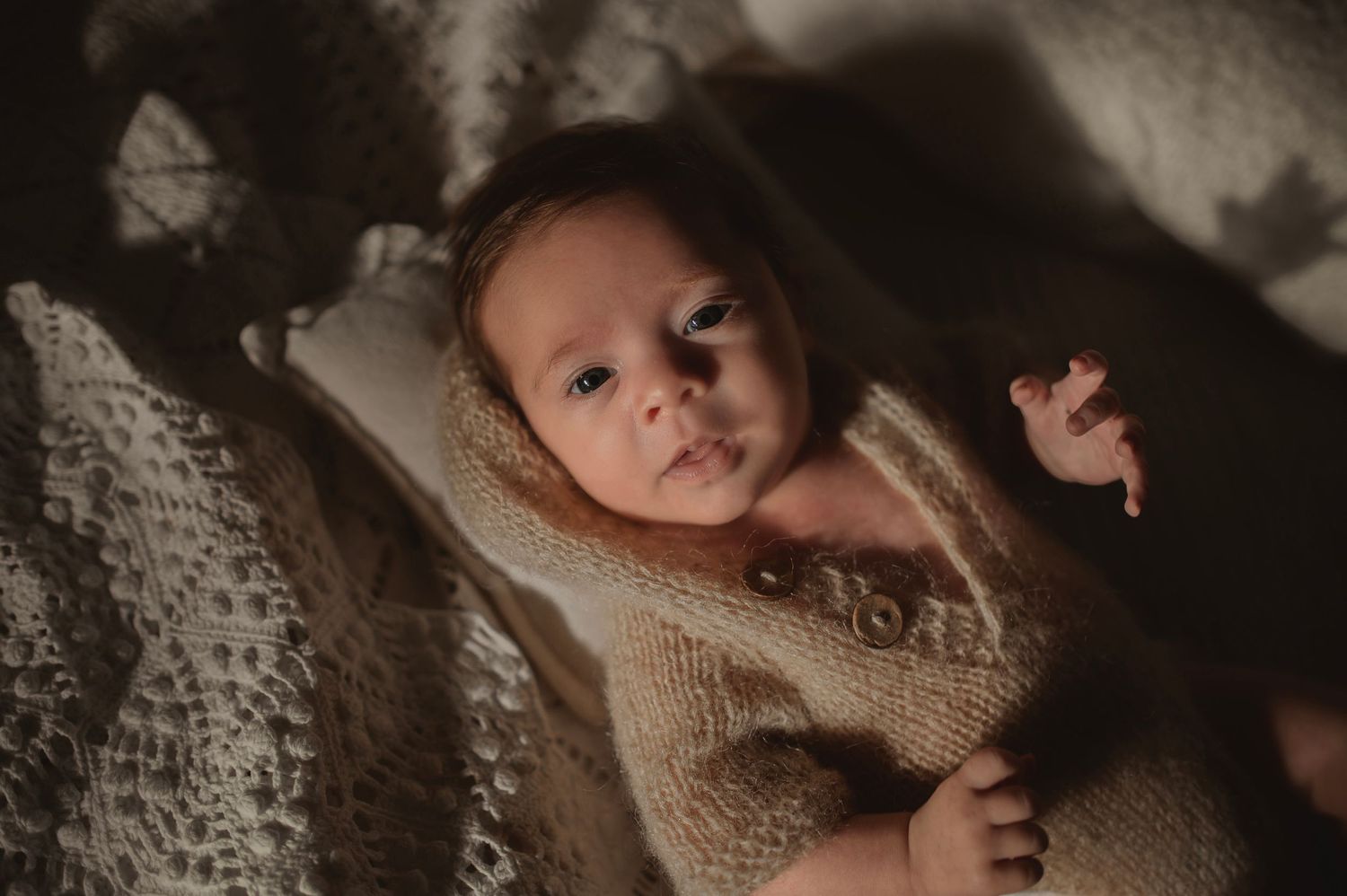 Baby in beige knit sweater lying on textured white bedding with moody lighting.