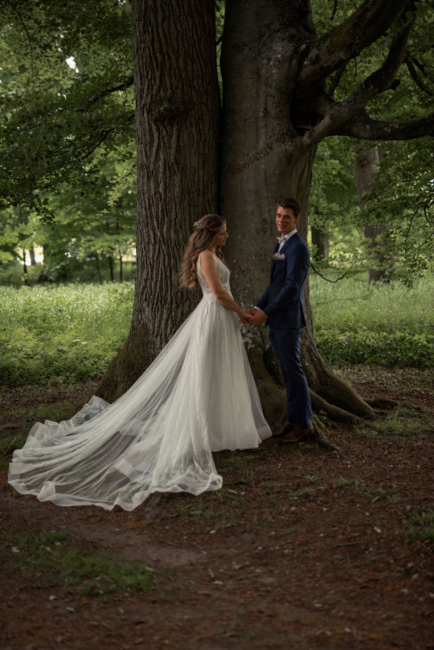 Elopement in Sweden with bride and groom holding hands under a large tree in a lush green forest, exchanging wedding vows.