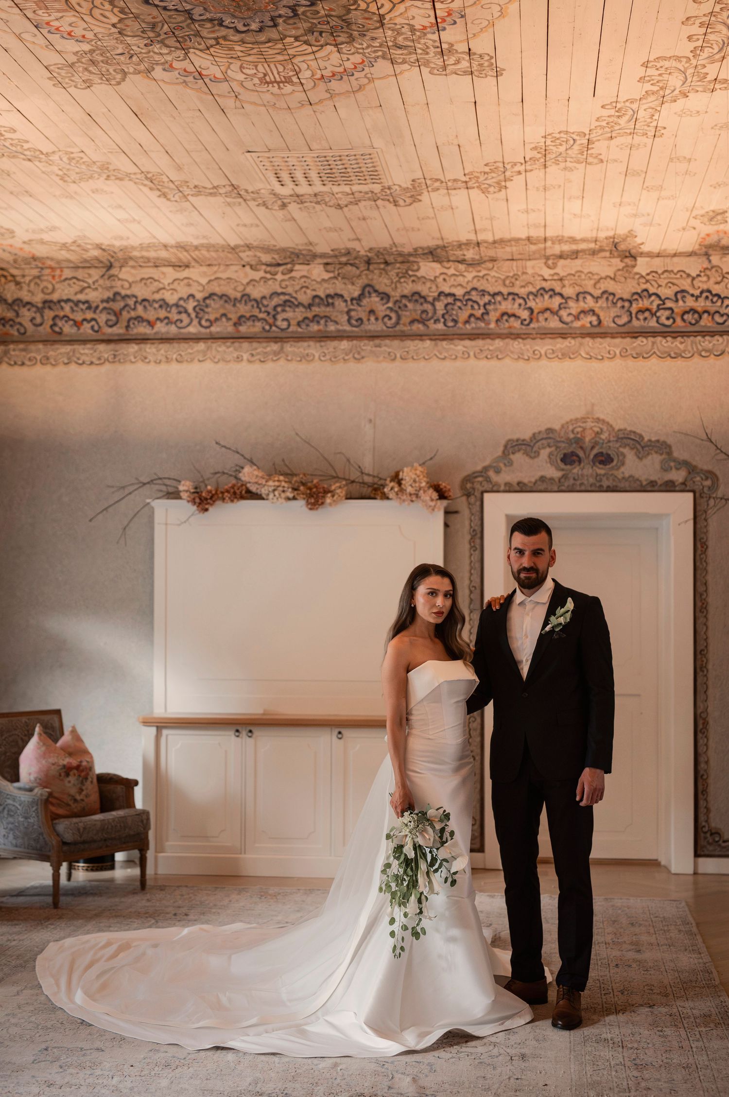 Brudpar fotograferat på Norrviken i Båstad med elegant interiör, målade tak och blomsterdekorationer i bakgrunden.