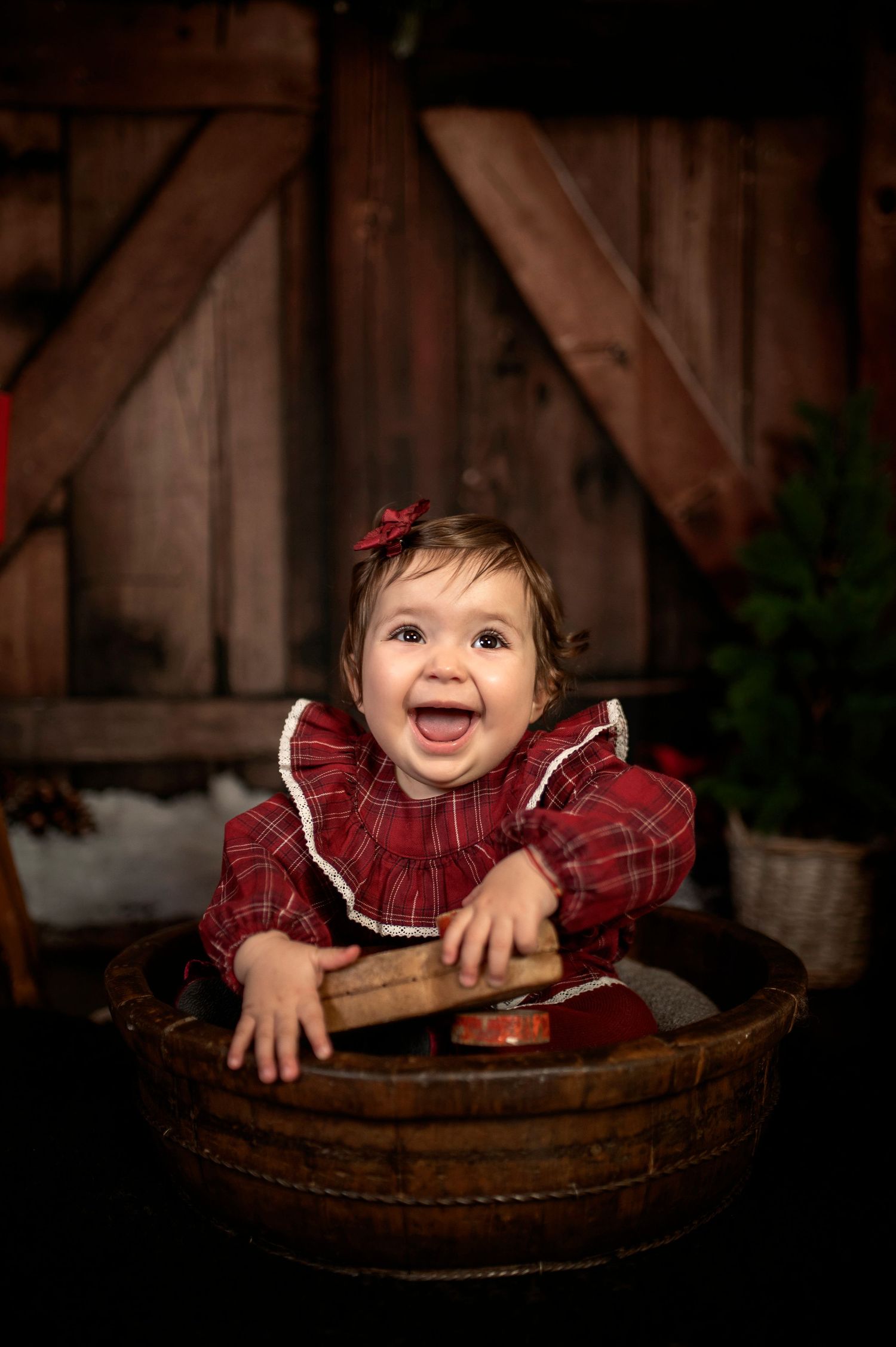 A joyful baby girl in a red plaid dress sits in a wooden tub, smiling brightly in a rustic barn setting.