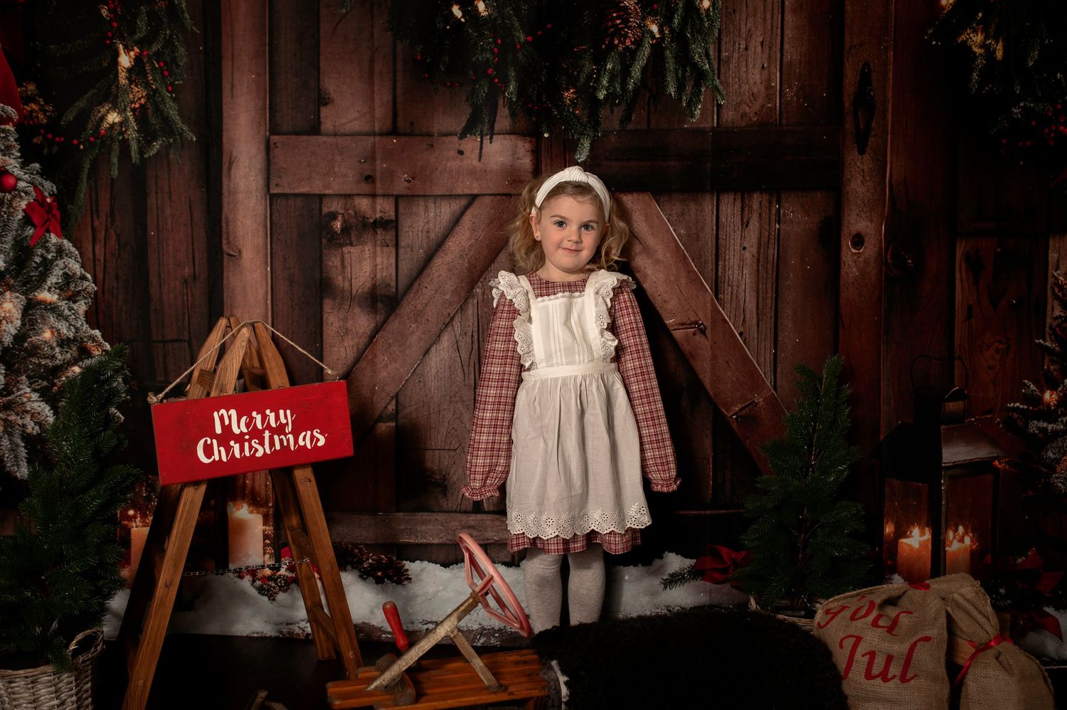 Young girl in plaid dress poses by Merry Christmas sign with festive holiday barn backdrop decor.
