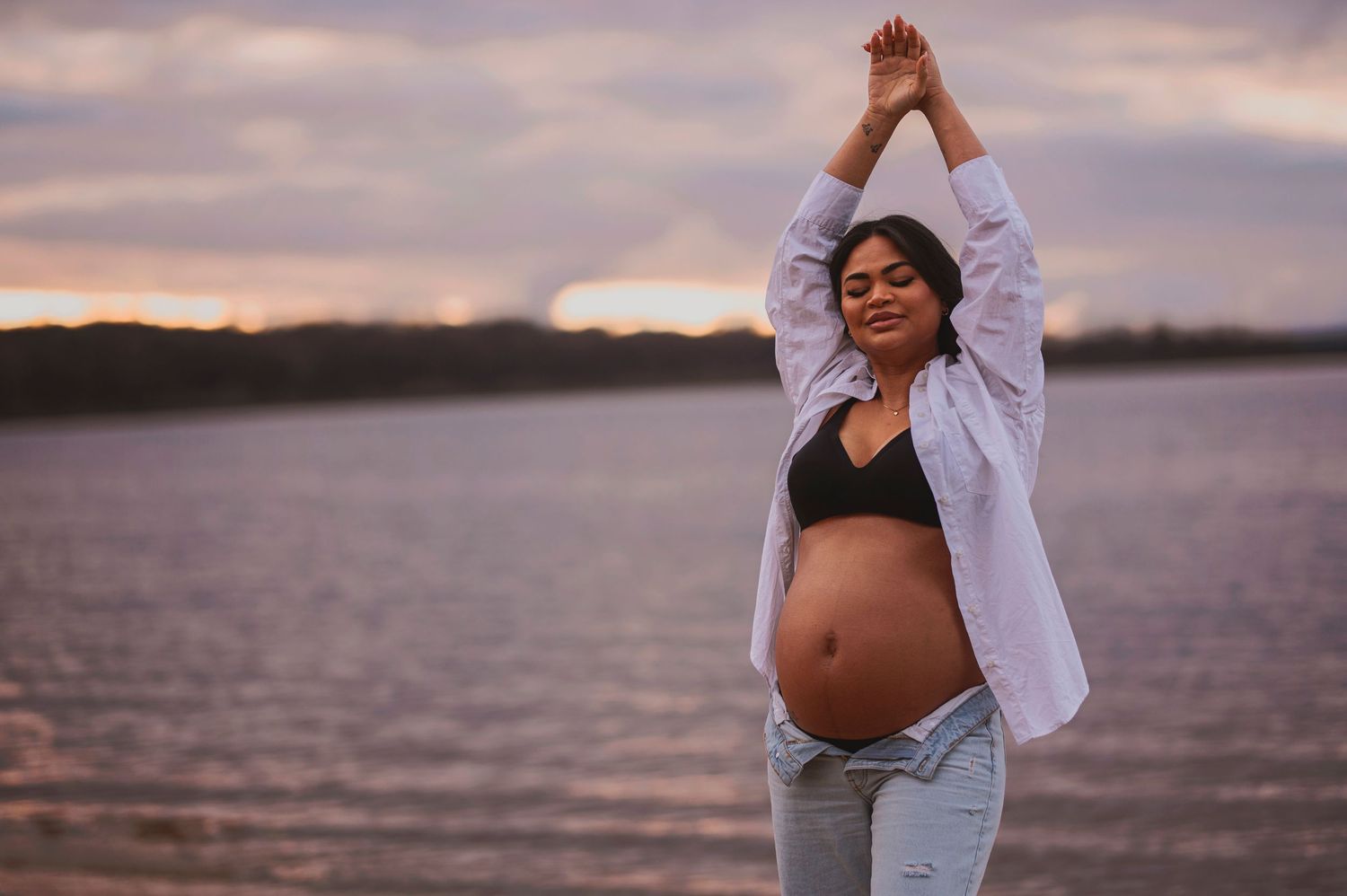 A pregnant person wearing a white shirt and jeans poses with arms raised by a tranquil lake at dusk.