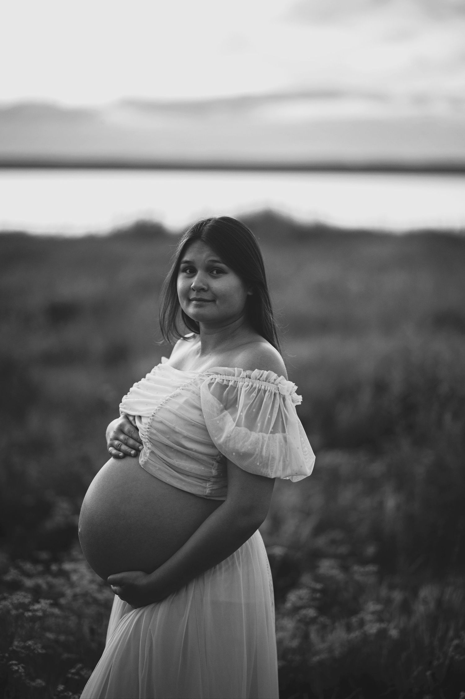 A black and white maternity portrait featuring an off-shoulder white dress against a scenic landscape.