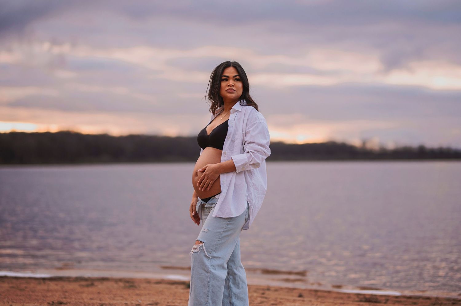 Expectant mother in white shirt and jeans stands contemplatively by lakeside at dusk.