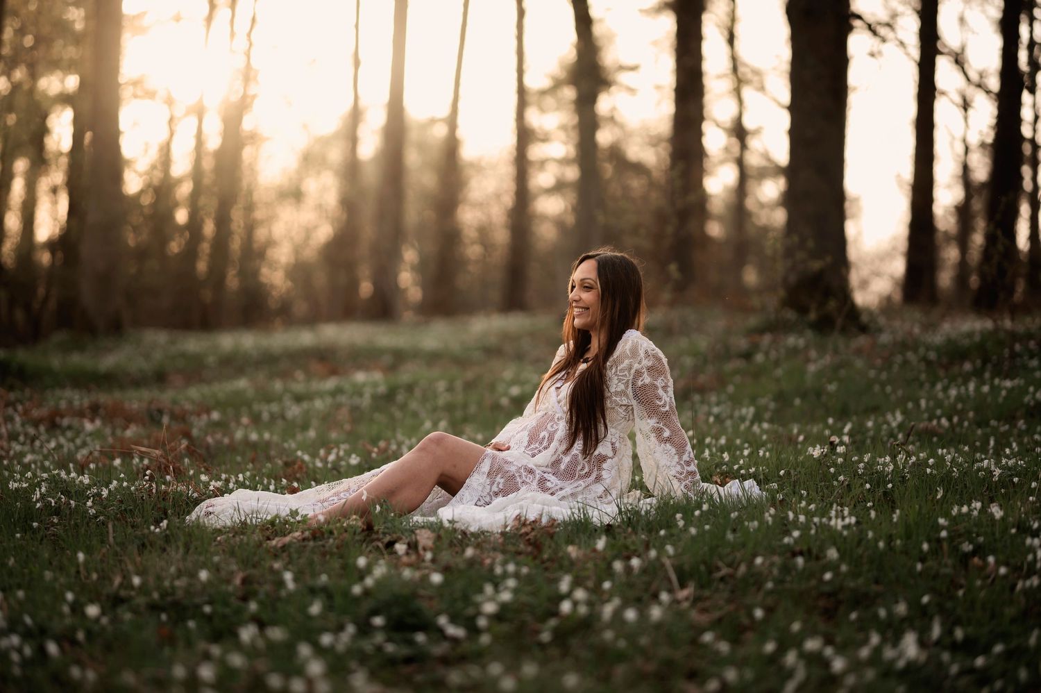 Pregnant woman in white lace dress sitting in a field of wildflowers at golden hour.