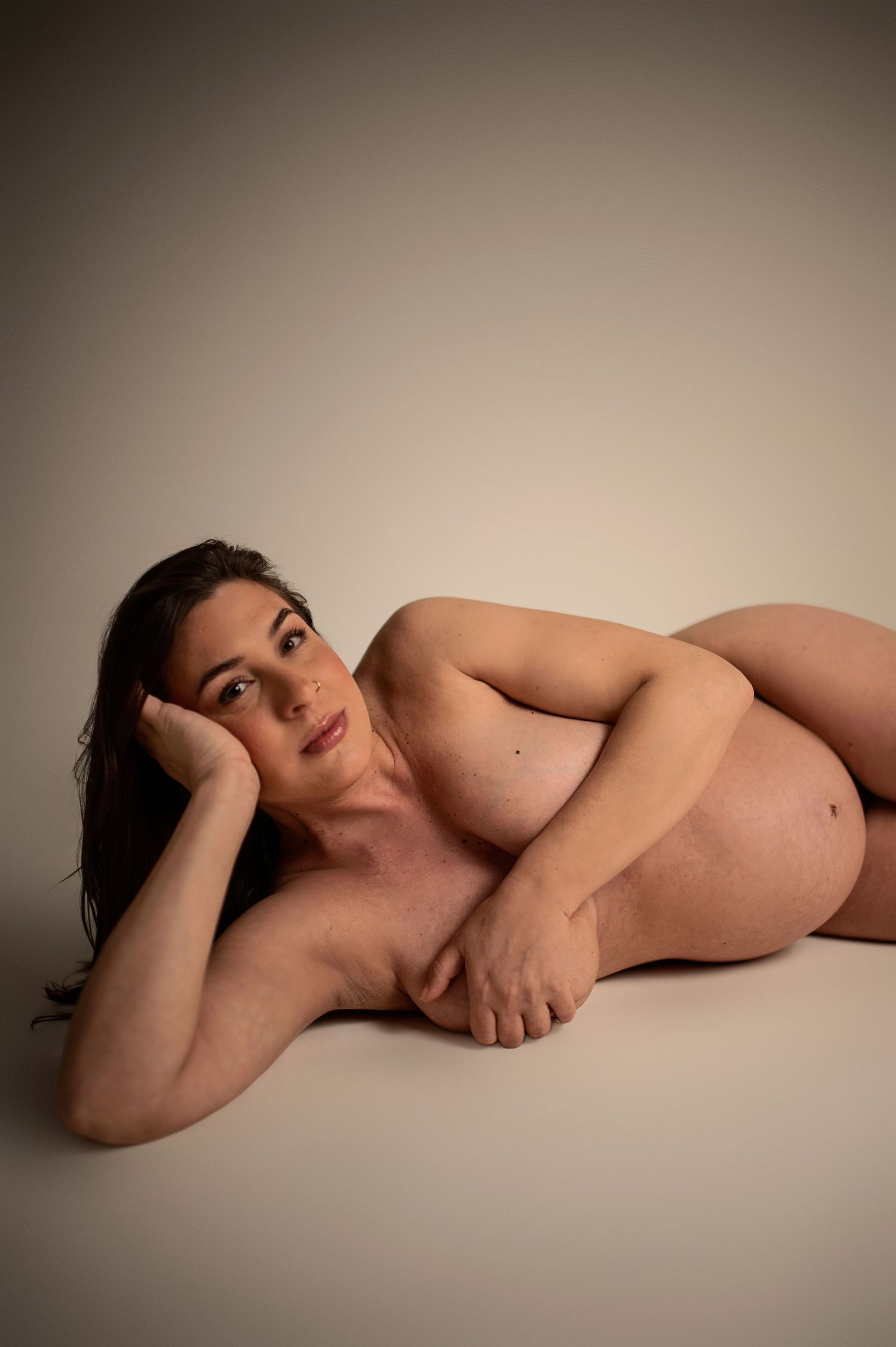 Nude pregnant woman lying on her side in a studio, resting her head on her hand against a neutral background.