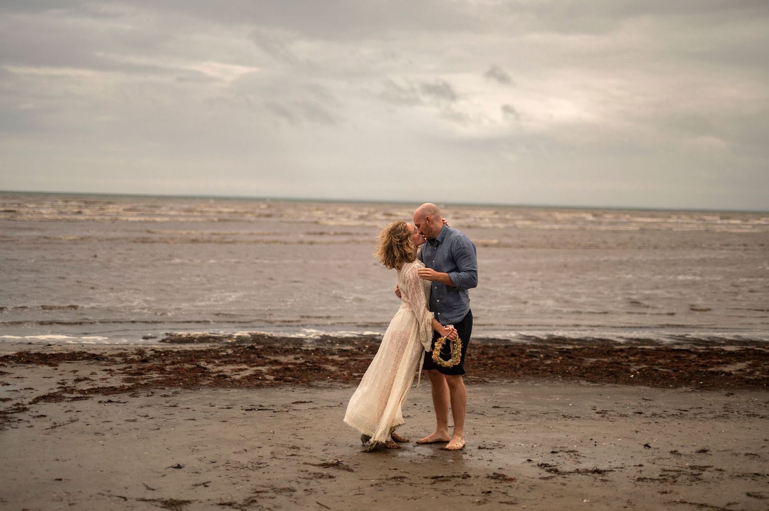 Wedding photography in Båstad, Sweden with barefoot couple sharing a romantic kiss on a cloudy beach with waves and wet sand.