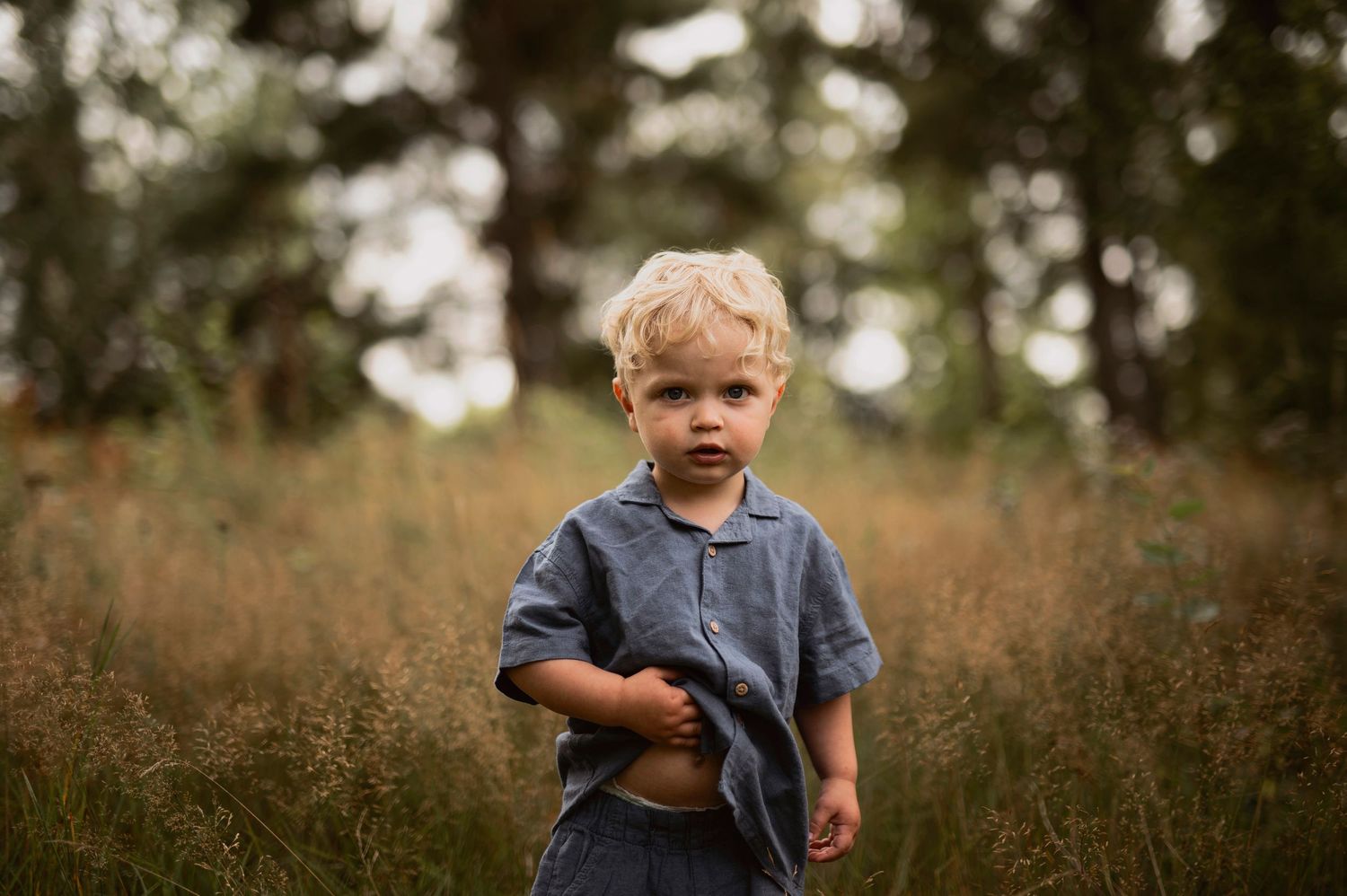Barnfotografering i Skåne med blond liten pojke i blå tröja i högt gräs med träd i bakgrunden.