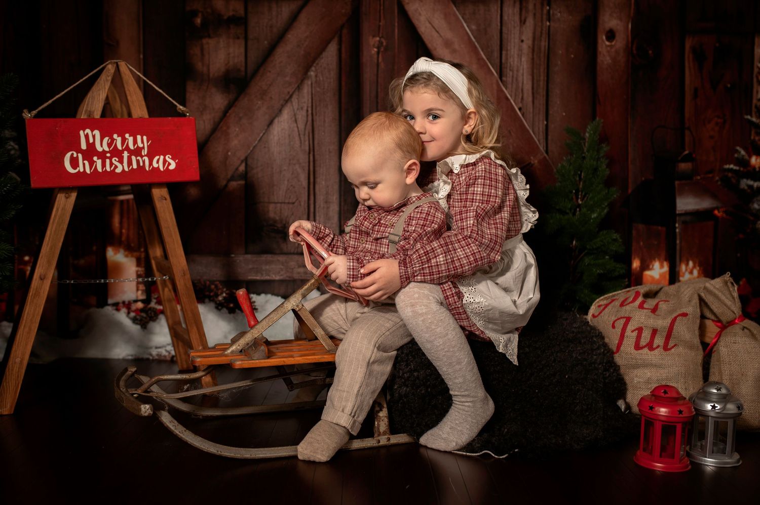 Two young children sit together on a vintage sled near a Merry Christmas sign in a festive holiday photo.