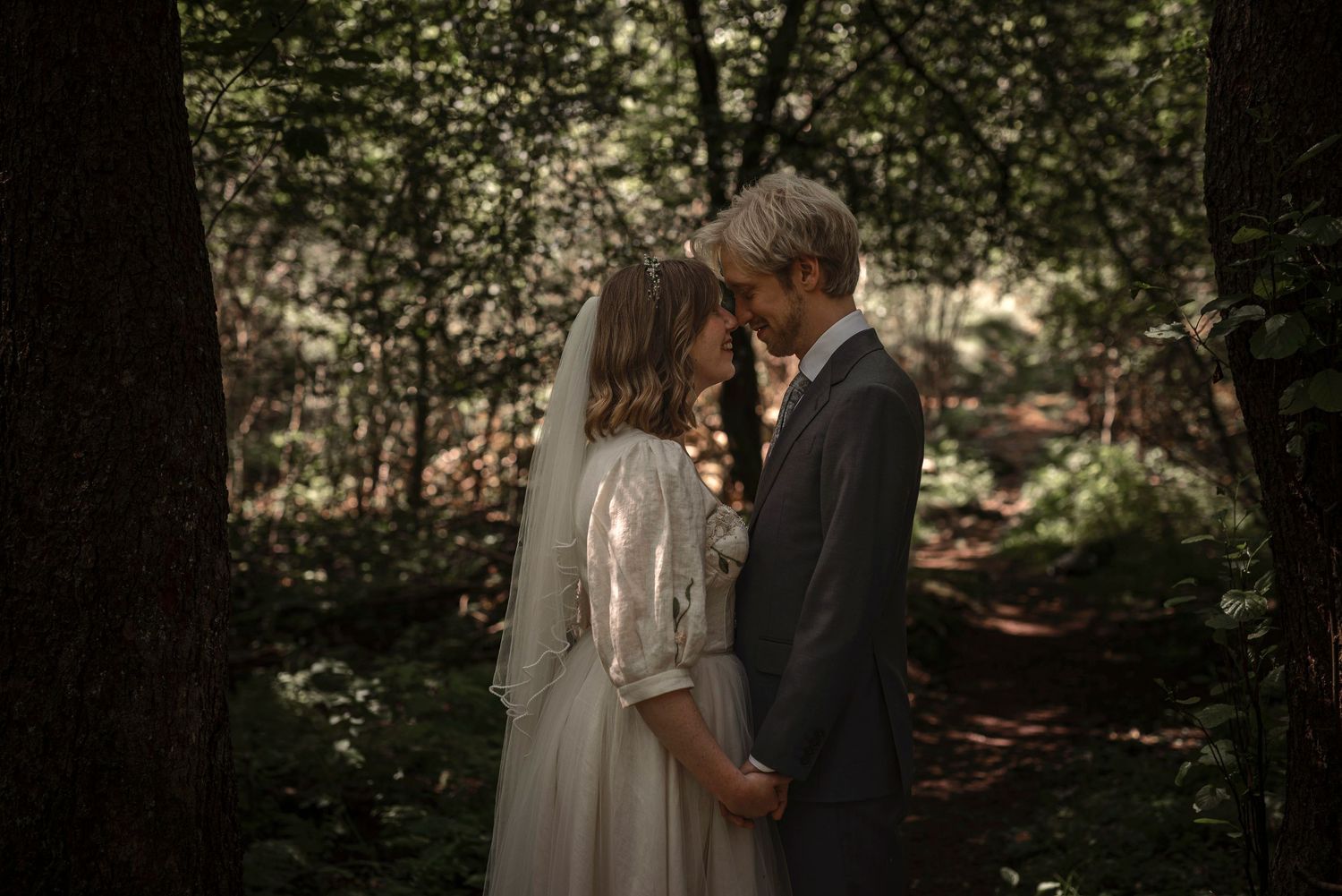 A bride and groom share an intimate moment in a lush forest, the bride wearing a white gown and veil.