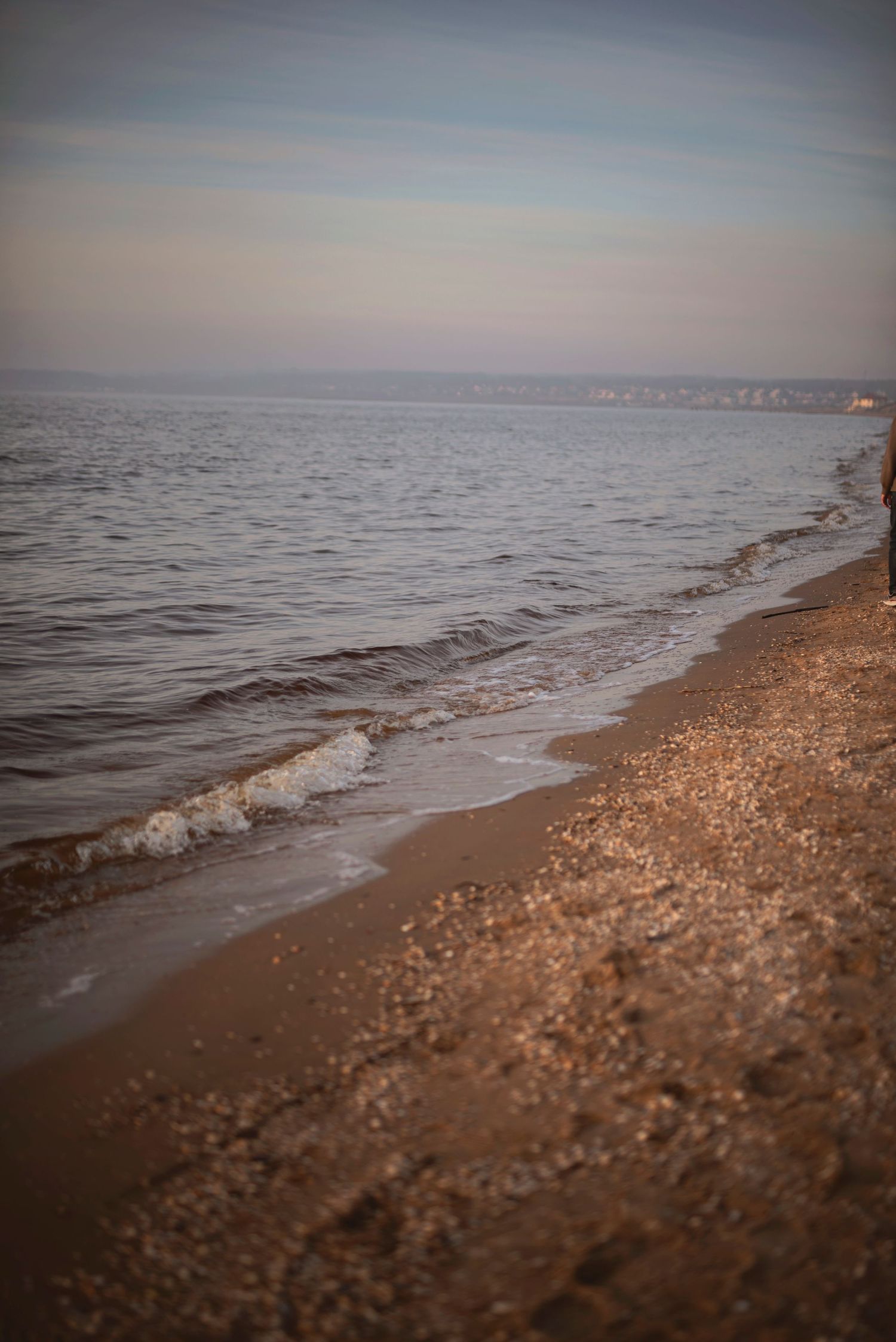 Gentle waves wash up on a sandy beach shoreline during a tranquil sunset with misty horizon in the distance.