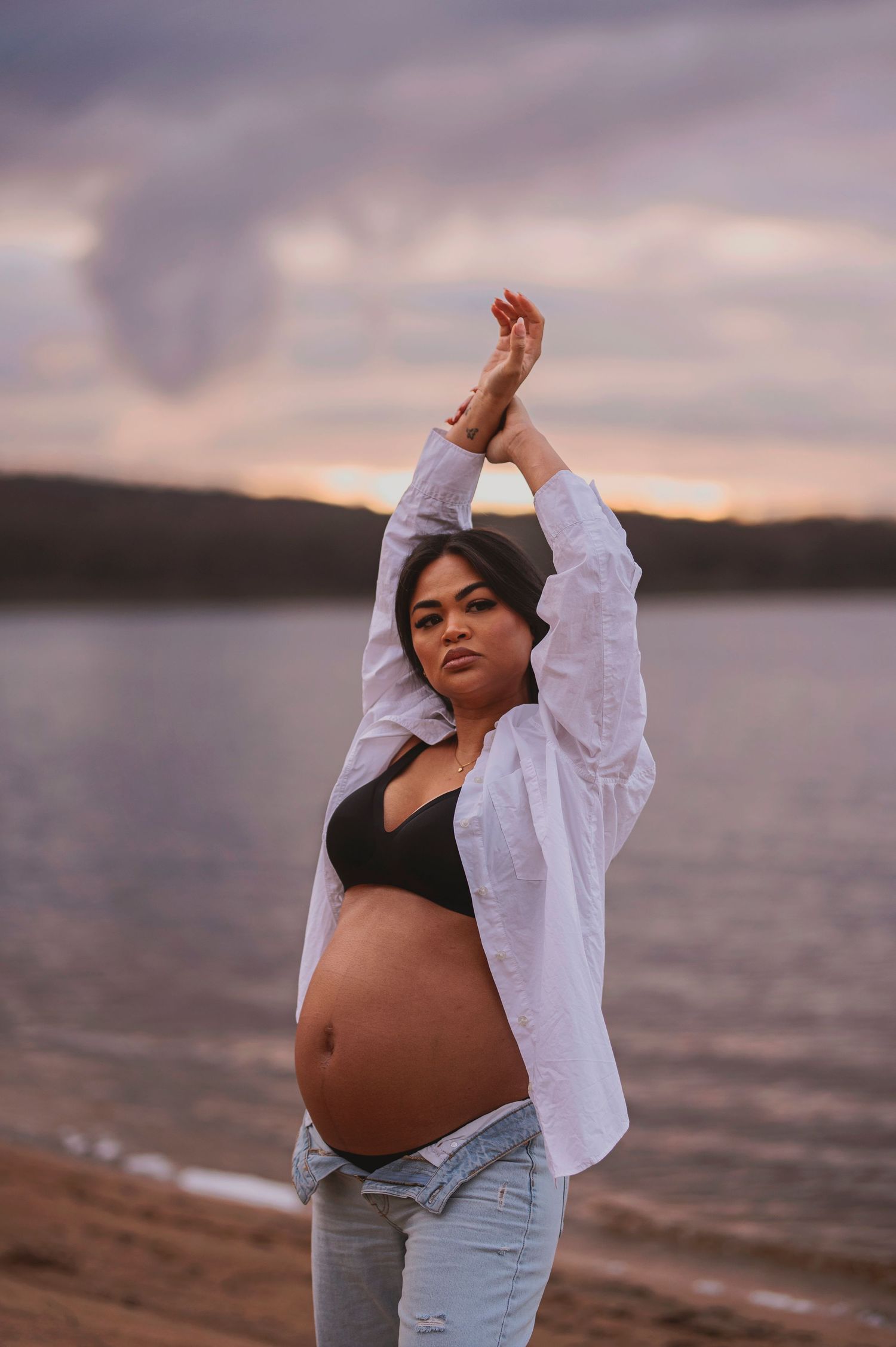 Maternity portrait with raised arms pose against sunset lake backdrop.