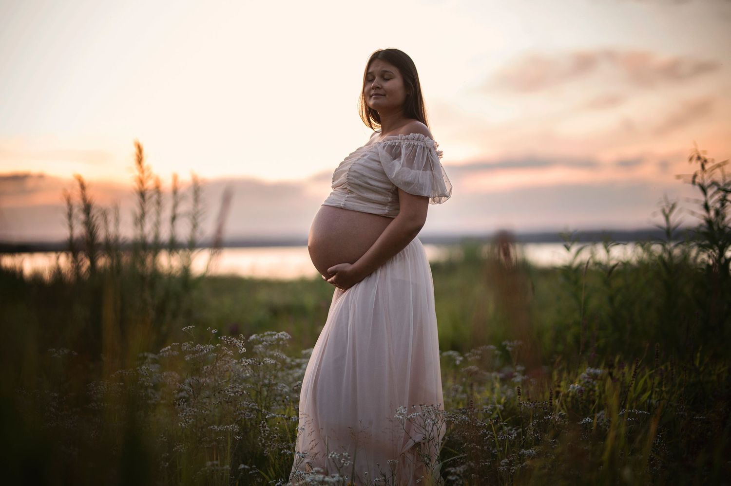 A maternity portrait in a white dress against a sunset backdrop near water.