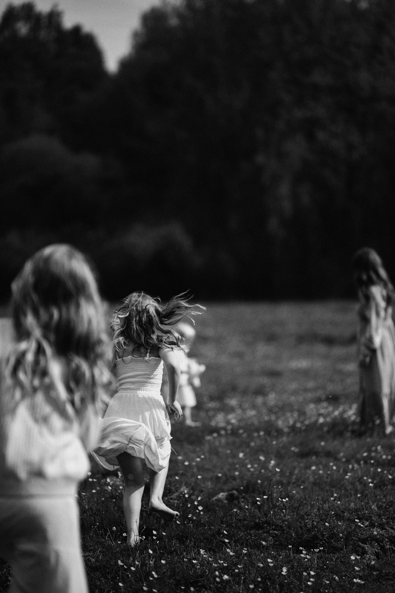 Familjefotografering i Skåne med flickor i vita klänningar som springer barfota i äng i svartvitt ljus.
