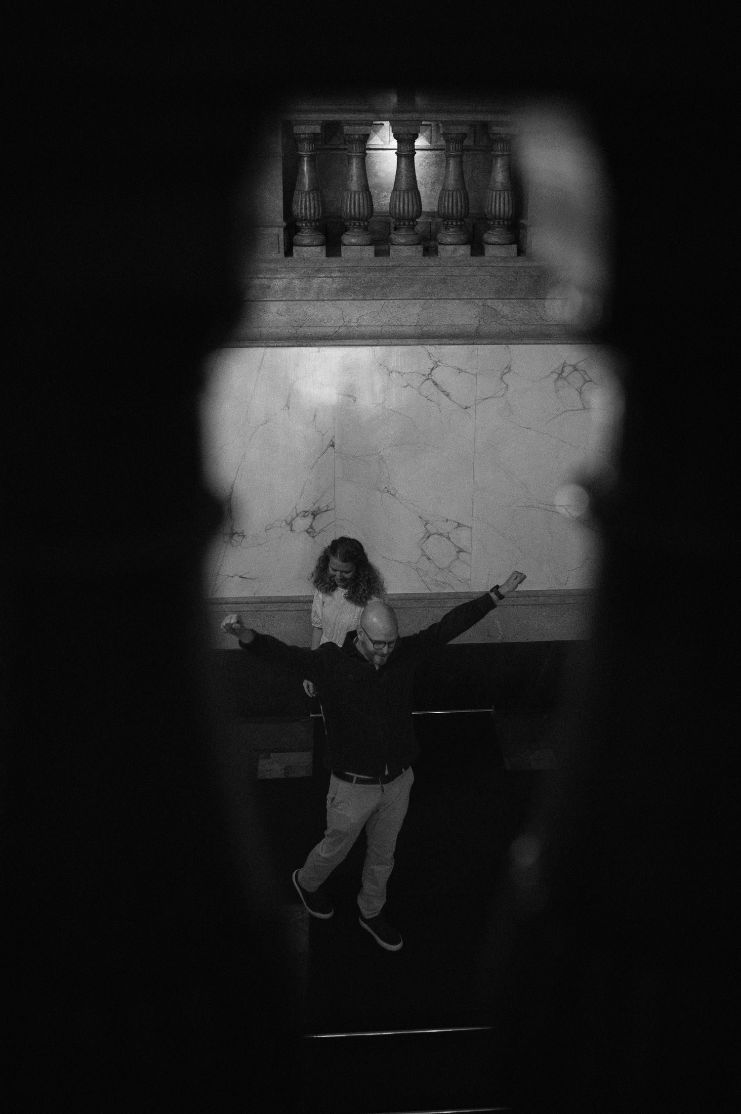 Black and white artistic photo of couple dancing in grand marble hall viewed through dark archway frame.