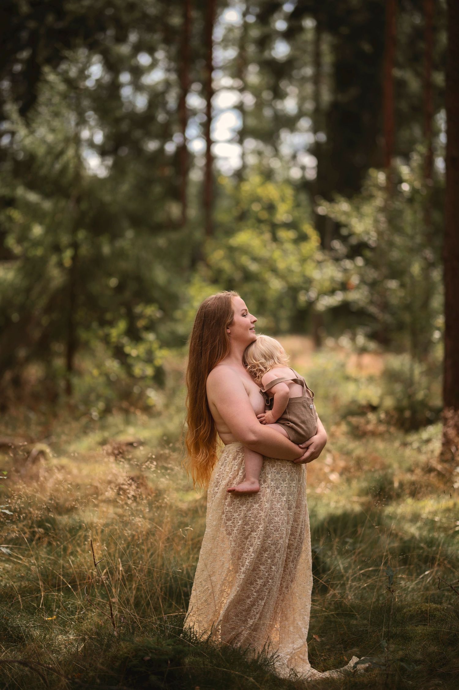 Moderskapsfotografering i Skåne med förälder som håller ett litet barn i spetsklänning i drömlik skogsmiljö.