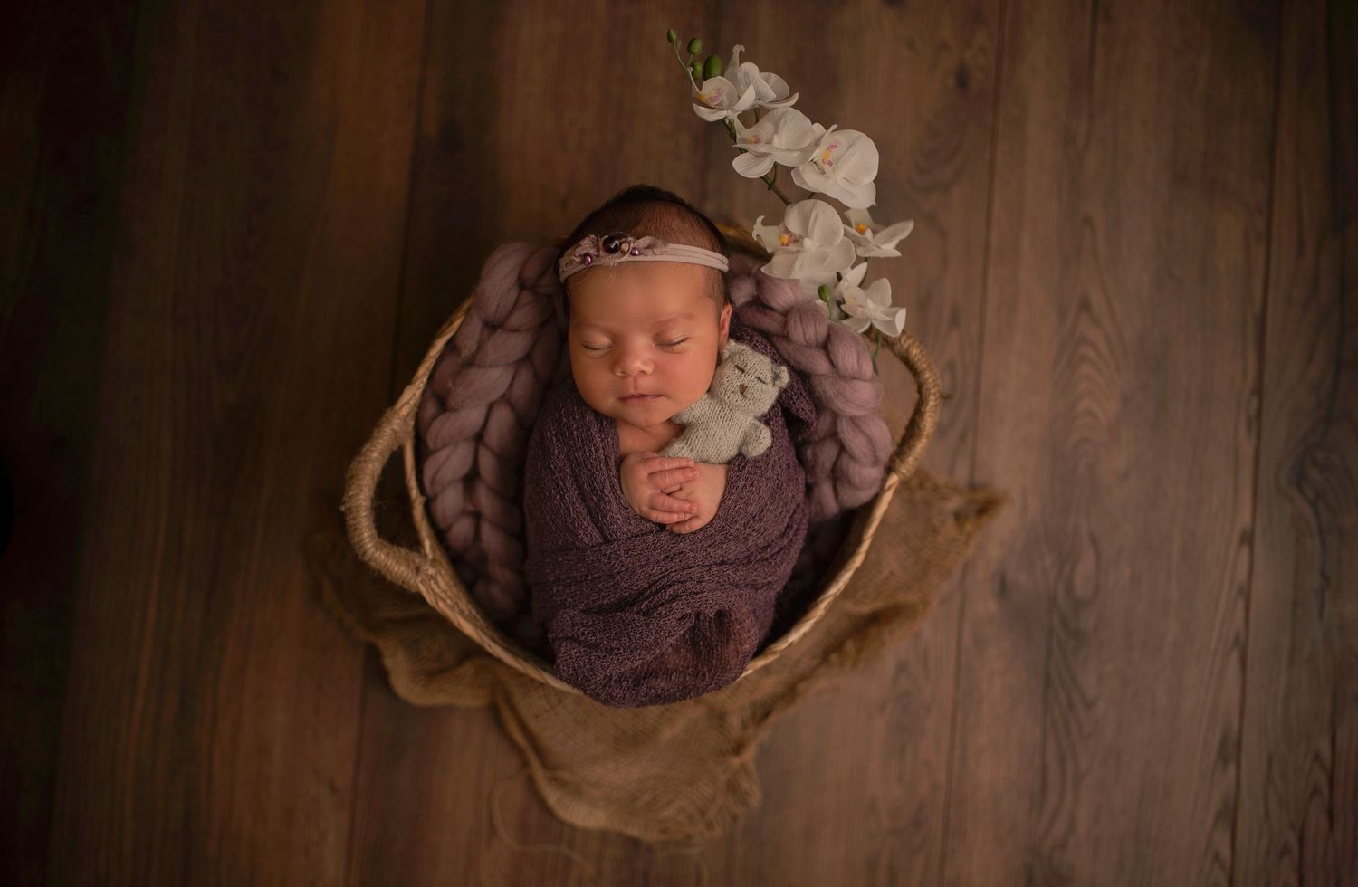 Newborn sleeping in wooden bowl with purple flowers and brown fabric.