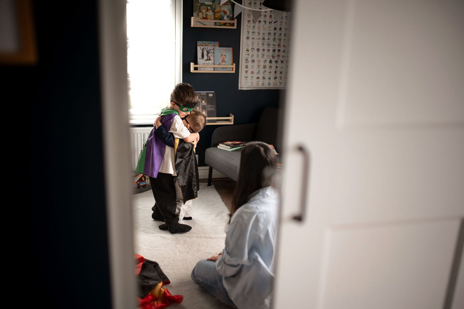 Child in colorful costume stands in bedroom while adult sits on floor, viewed through partially open door.