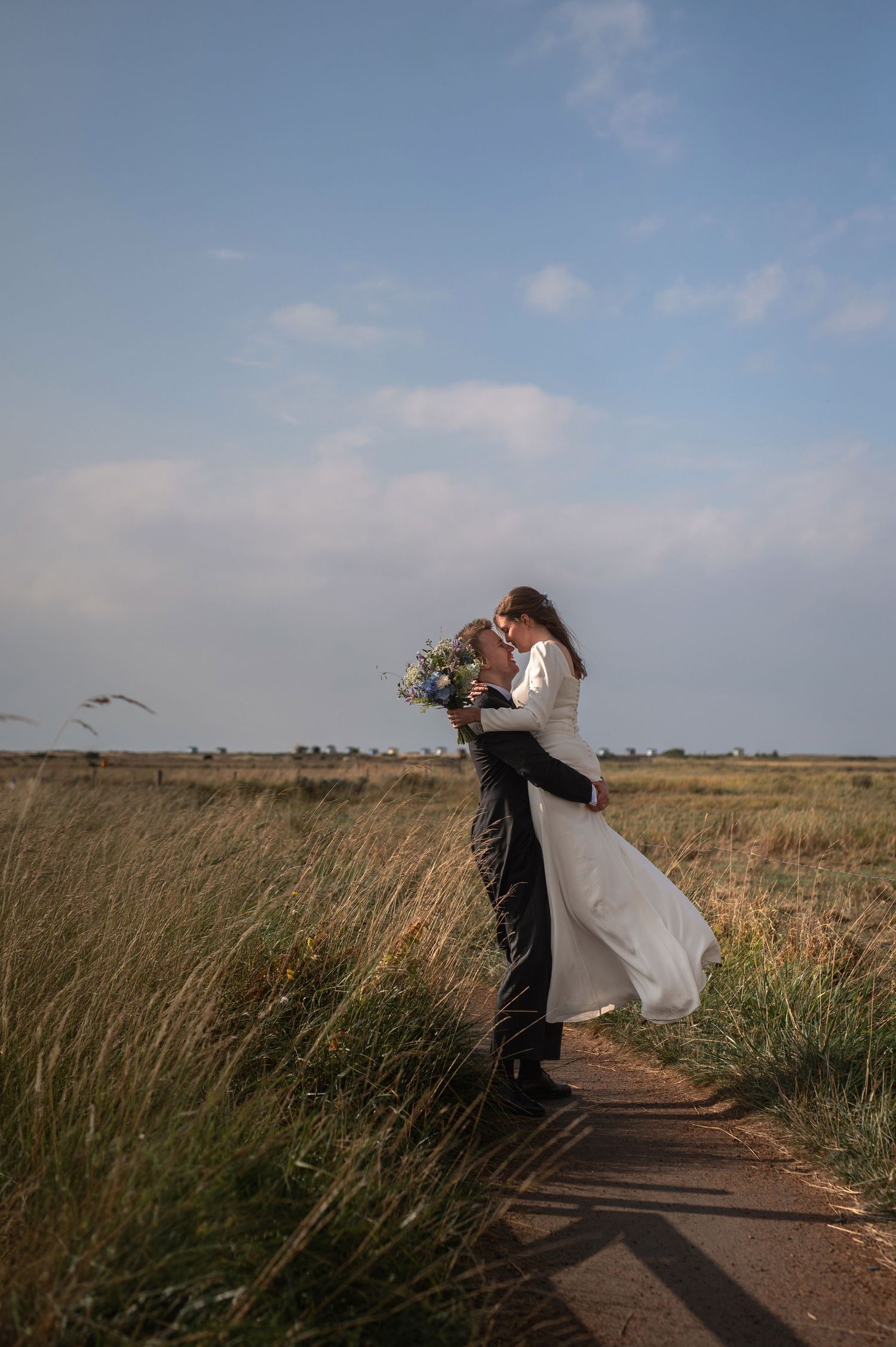 Groom lifting bride in white dress on a rural path, surrounded by golden fields under a blue sky.