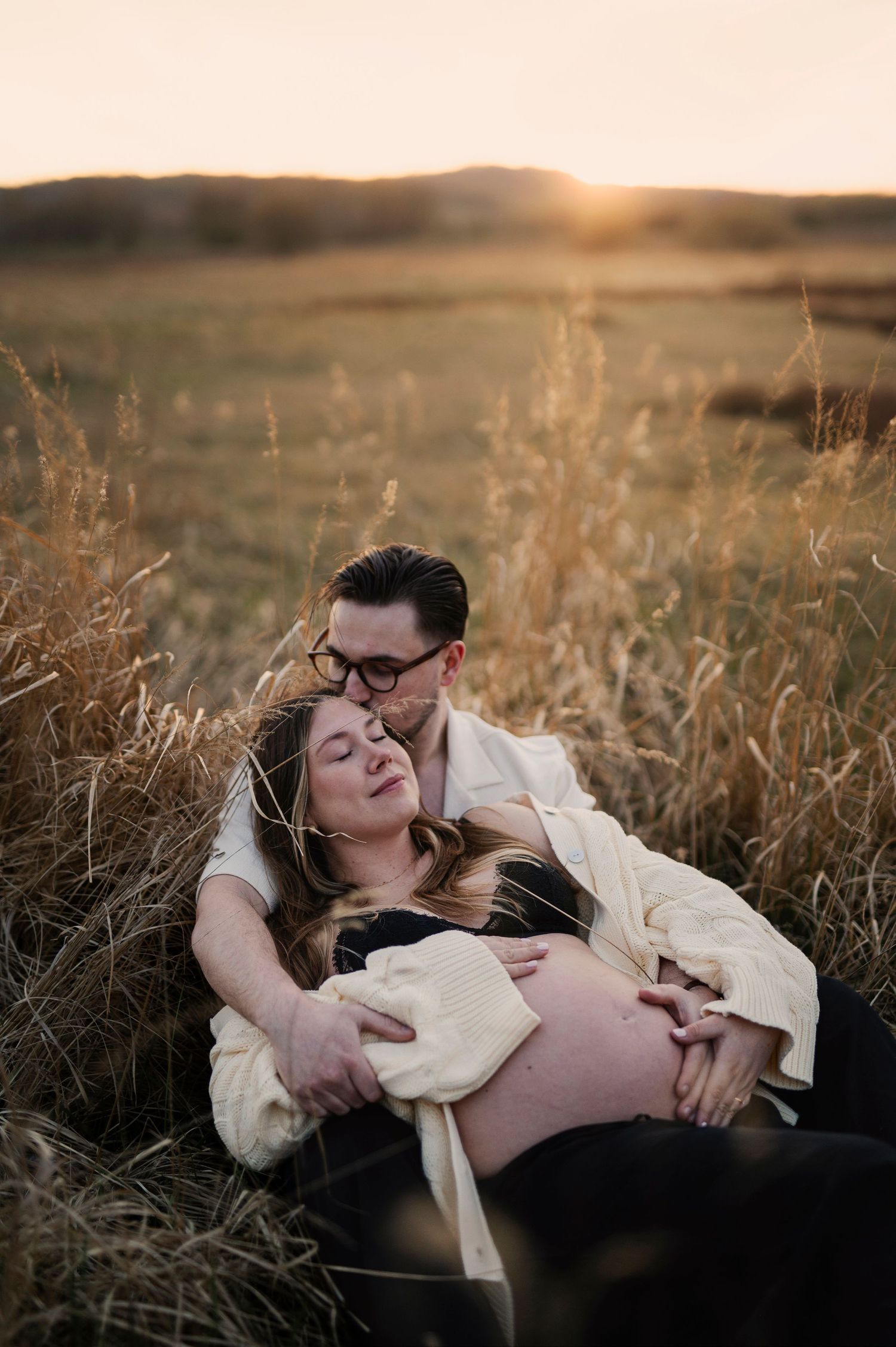 Couple sitting in golden field at sunset, man embracing pregnant woman from behind in tender maternity photo.