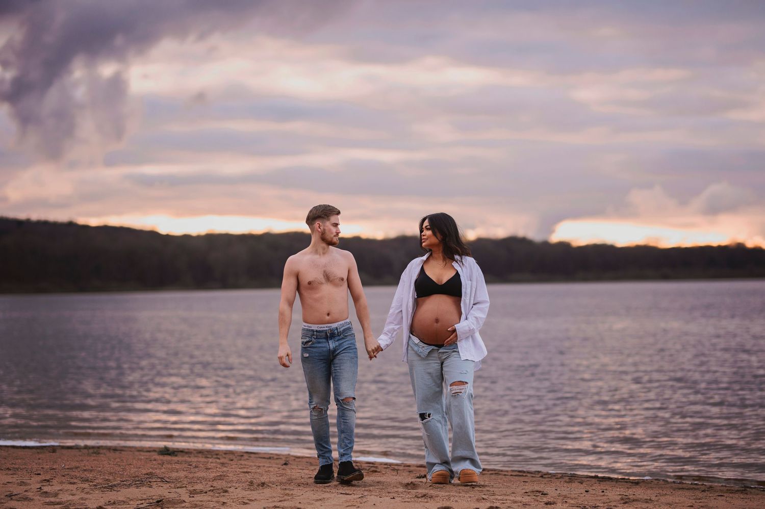 A couple stands together on a beach at sunset with a lake and wooded shoreline in the background.