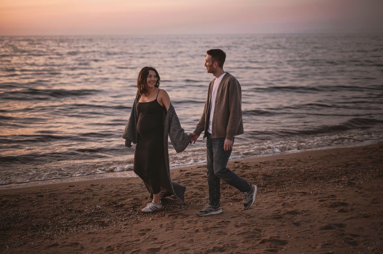 A couple holds hands while walking along a beach at sunset with the ocean waves rolling in behind them.