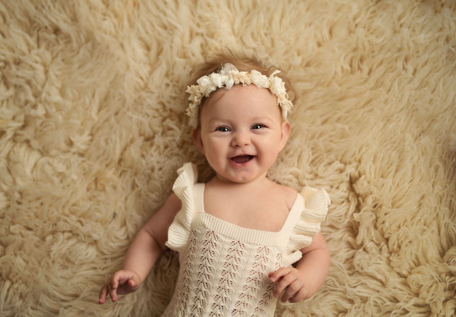 Baby wearing floral crown and knit outfit smiles on cream fur background.
