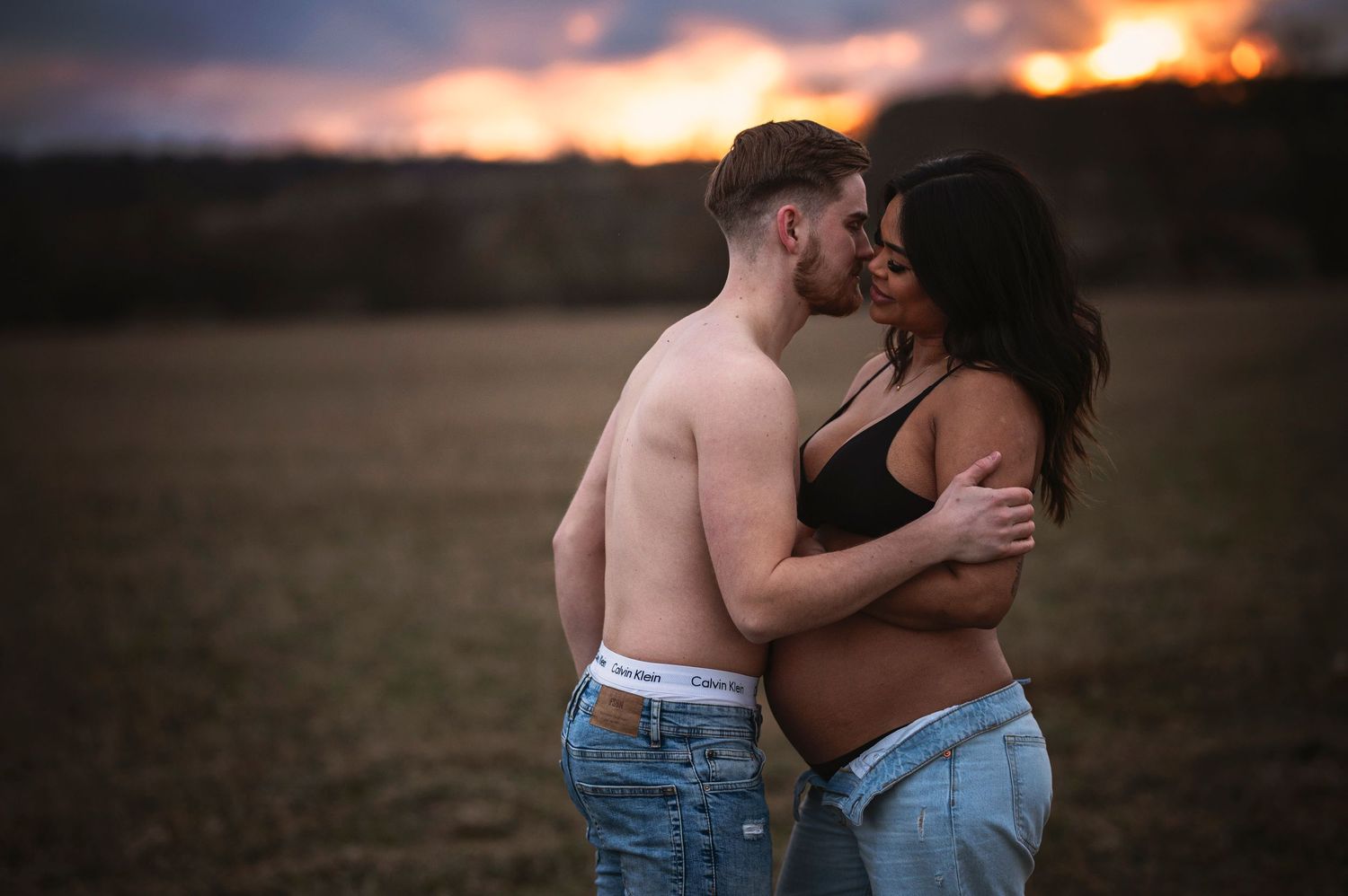 An expecting couple shares an intimate moment in a field during golden hour.