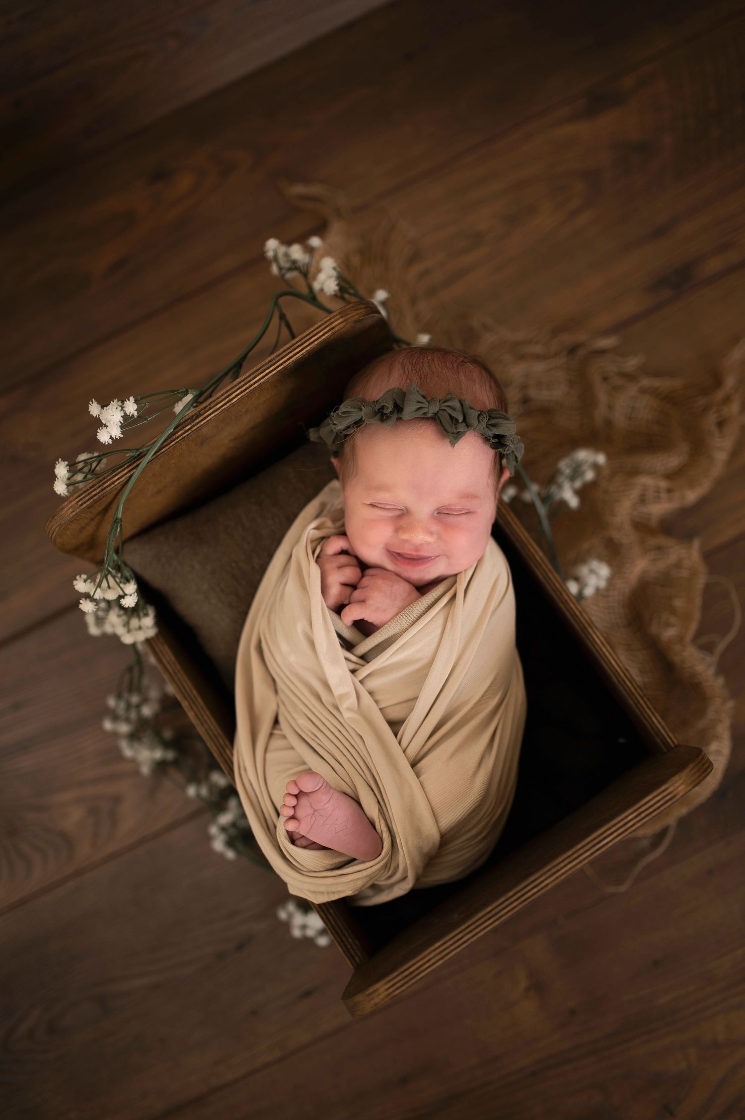 Newborn baby wrapped in beige swaddle sleeping in wooden box with delicate flowers.
